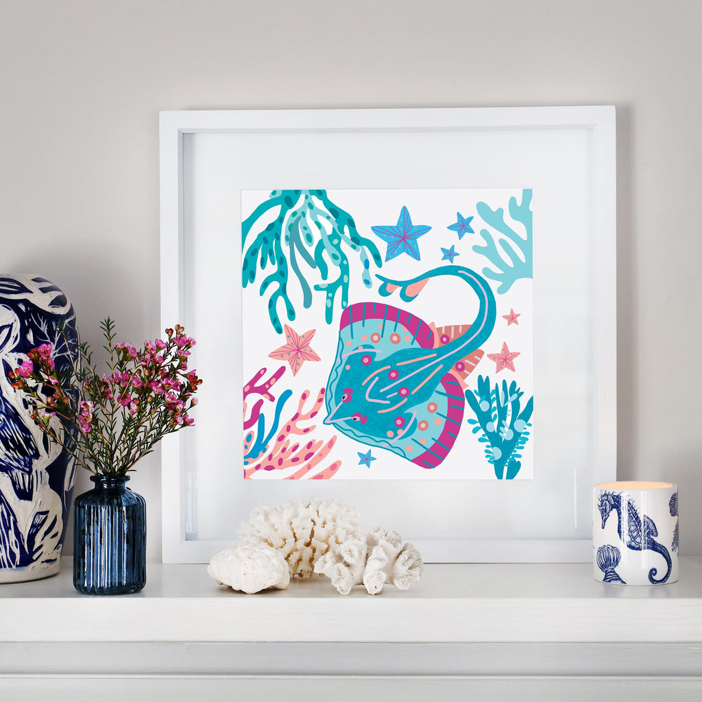 A framed illustration of a colorful skate swimming among seaweed, starfish, and coral in shades of blue, pink, and turquoise, displayed on a white mantel. The scene is decorated with a blue glass vase holding pink flowers, a white coral piece, a ceramic cup with a seahorse design, and a navy and white patterned sculpture.