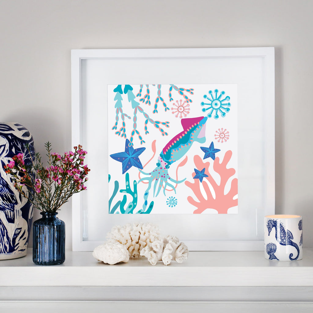 A framed illustration of a vibrant squid swimming among starfish, coral, and seaweed in shades of blue, pink, and turquoise, displayed on a white mantel. The decor includes a blue glass vase with pink flowers, white coral pieces, a ceramic cup with a seahorse design, and a navy and white patterned sculpture.