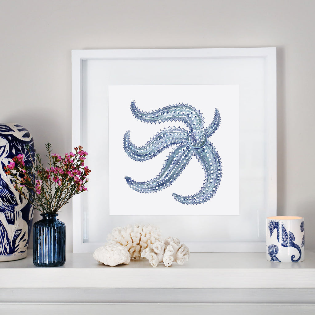 A coastal-inspired mantel display featuring a framed artwork of a detailed blue starfish. The arrangement includes a small blue glass vase with pink flowers, white coral pieces, a blue and white ceramic sculpture, and a seahorse-patterned candle. The decor blends oceanic elements with soft, natural tones for a serene aesthetic.