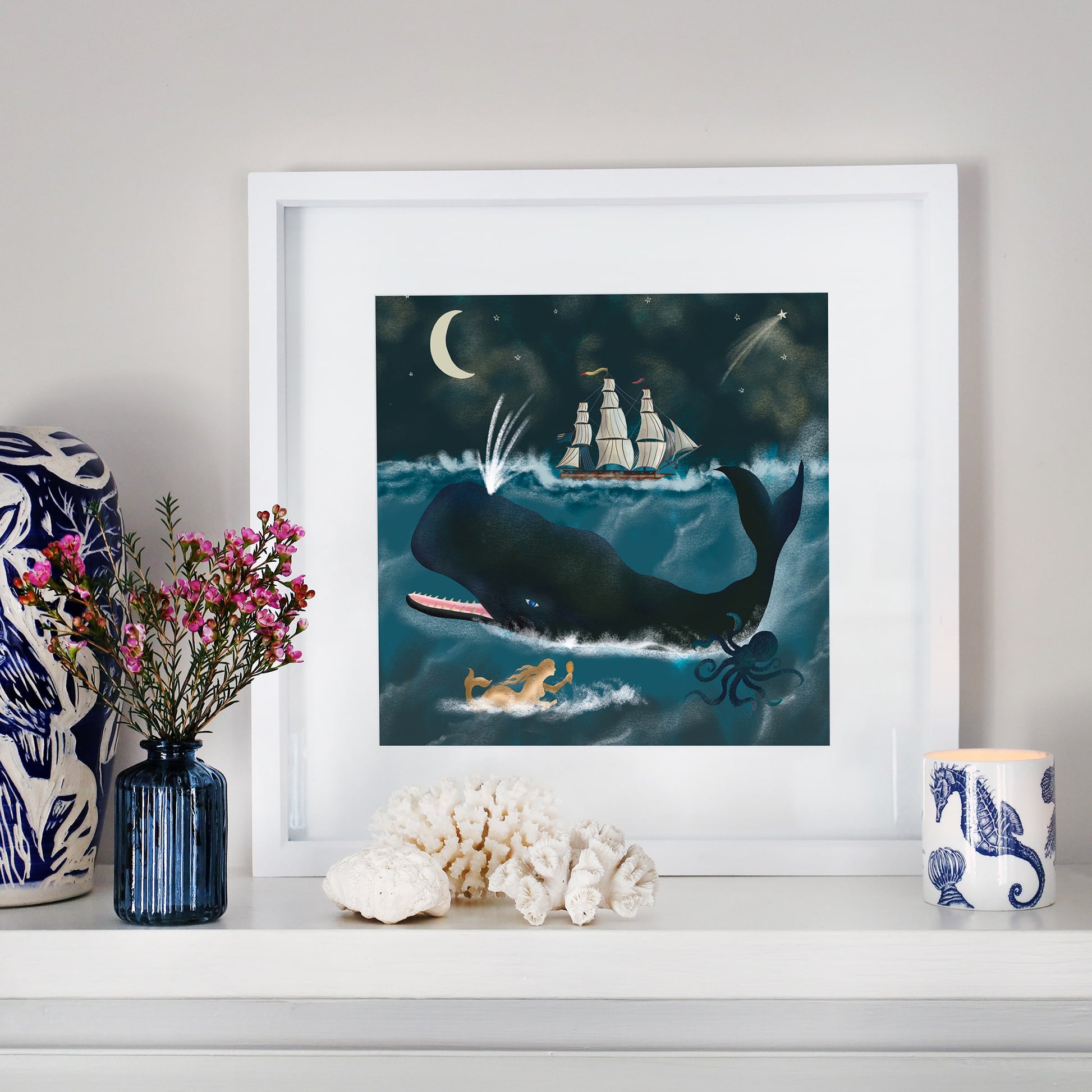 A framed ocean-themed illustration on a white mantel. The artwork depicts a large whale spouting water beneath a crescent moon, with a sailing ship on the horizon, a mermaid swimming, and an octopus below the waves. The mantel is decorated with coral, a blue glass vase with pink flowers, a ceramic candle holder with a seahorse design, and a blue and white patterned sculpture.