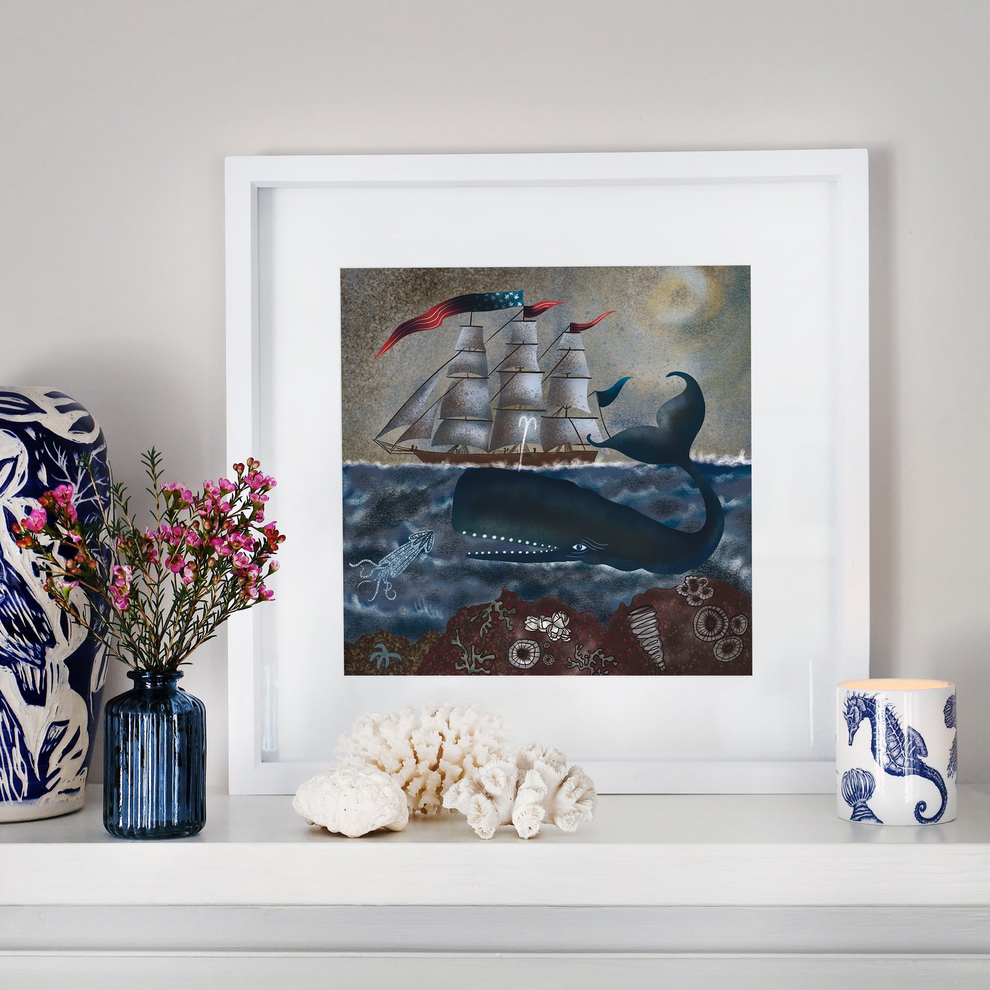 A framed nautical illustration of a whale and a sailing ship under a moonlit sky, placed on a white mantel. The scene includes coral, seashells, and a squid. Surrounding decor features a blue glass vase with pink flowers, white coral, a seahorse-patterned candle, and a ceramic piece with blue designs.