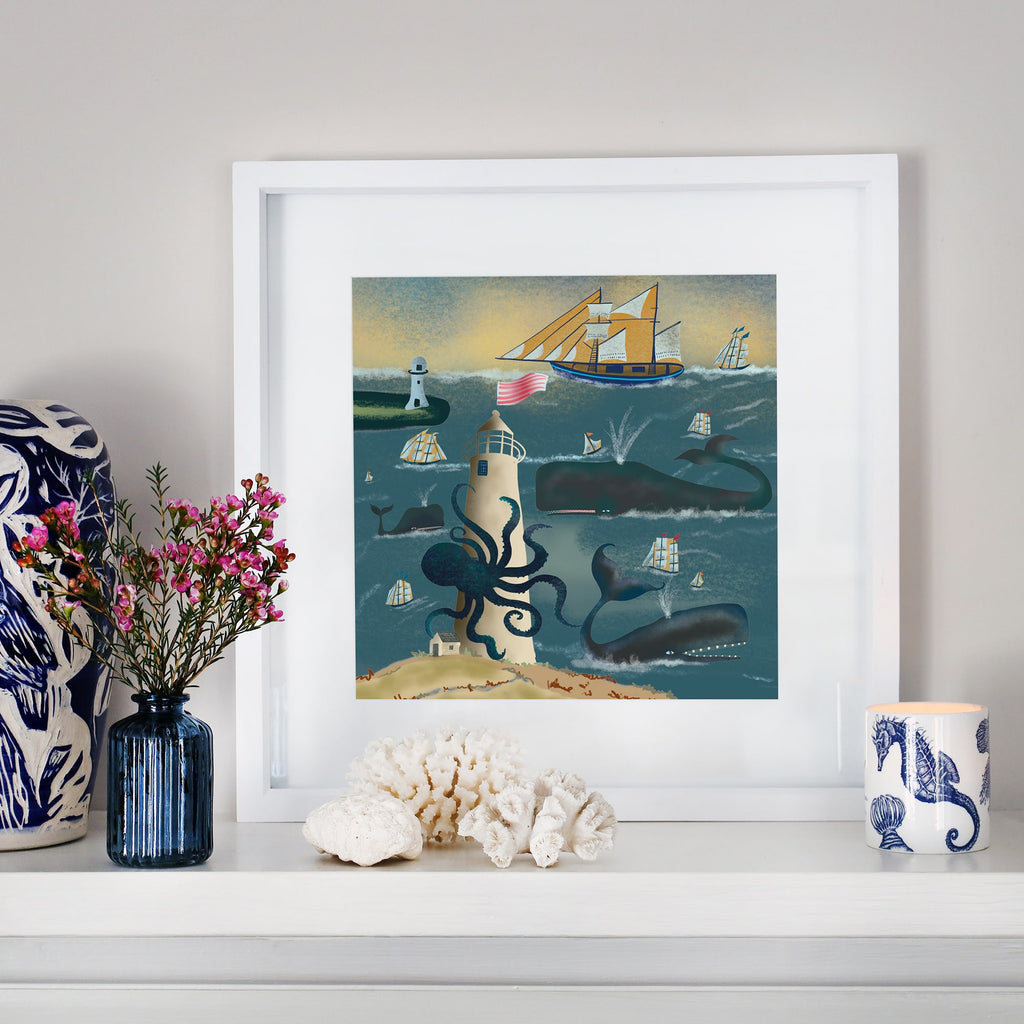 A framed nautical illustration displayed on a white mantelpiece. The artwork depicts a towering lighthouse entwined by a giant octopus, surrounded by sailing ships, whales, and crashing waves under a golden sky. The mantel is adorned with coral, a blue glass vase with pink flowers, a ceramic cup with seahorse patterns, and a decorative blue and white sculpture.