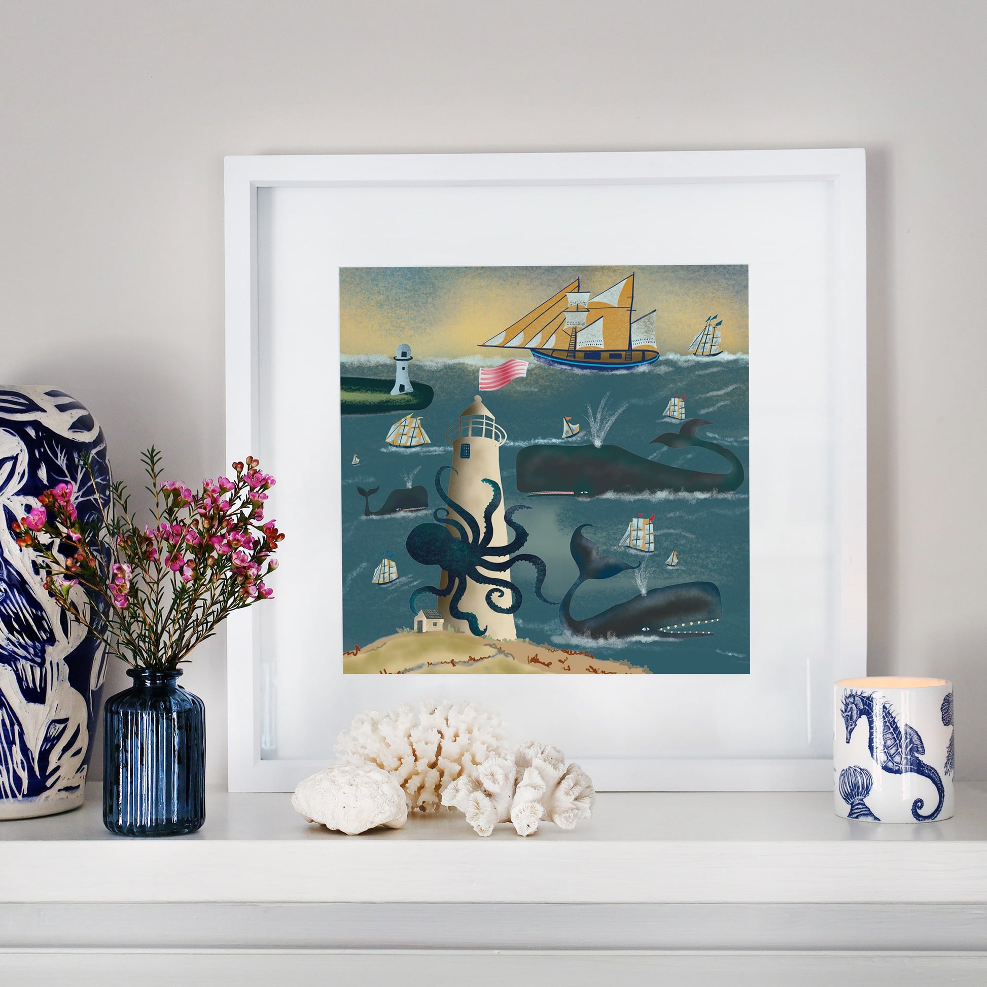 A framed nautical illustration displayed on a white mantelpiece. The artwork depicts a towering lighthouse entwined by a giant octopus, surrounded by sailing ships, whales, and crashing waves under a golden sky. The mantel is adorned with coral, a blue glass vase with pink flowers, a ceramic cup with seahorse patterns, and a decorative blue and white sculpture.