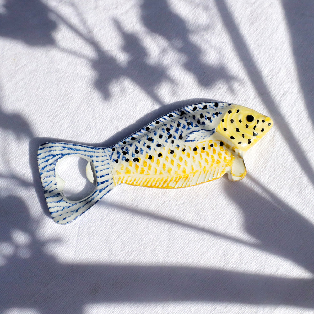 Small ceramic fish-shaped bottle opener with a yellow and blue patterned body, black spots, and a metal opener in the tail, placed on a white fabric background with soft shadows.