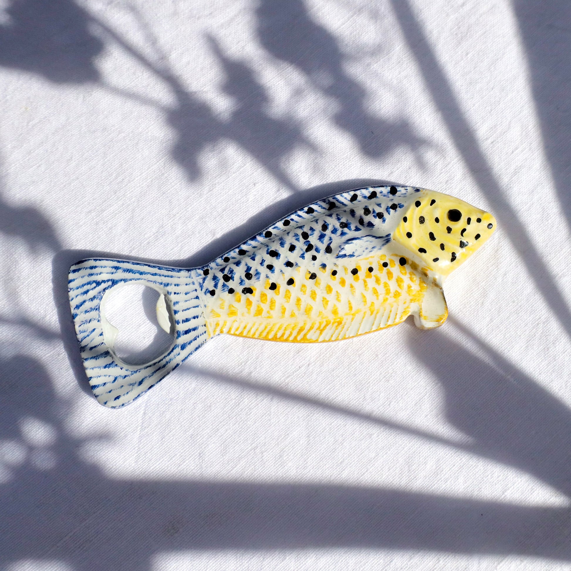 Small ceramic fish-shaped bottle opener with a yellow and blue patterned body, black spots, and a metal opener in the tail, placed on a white fabric background with soft shadows.