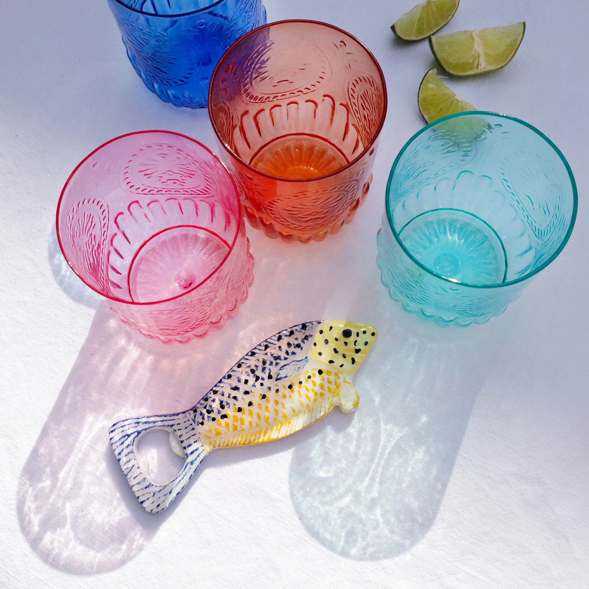 Colourful glass tumblers with a bottle opener and lime wedges on a white surface