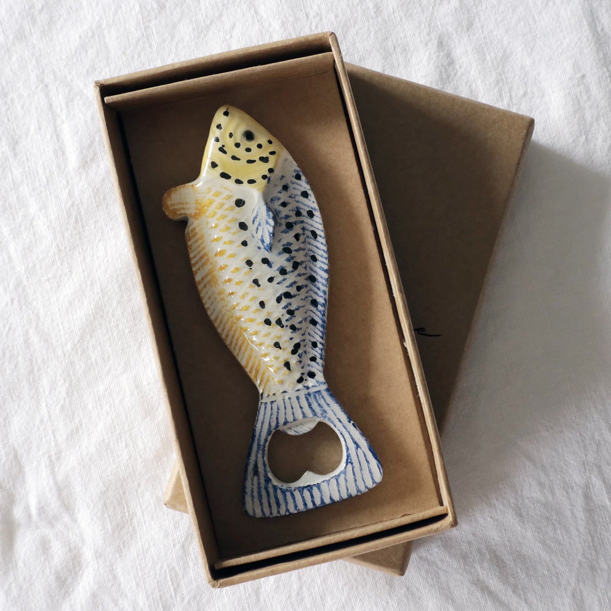 Fish-shaped bottle opener with yellow, blue, and black detailing, presented in a brown cardboard gift box on a white fabric background.