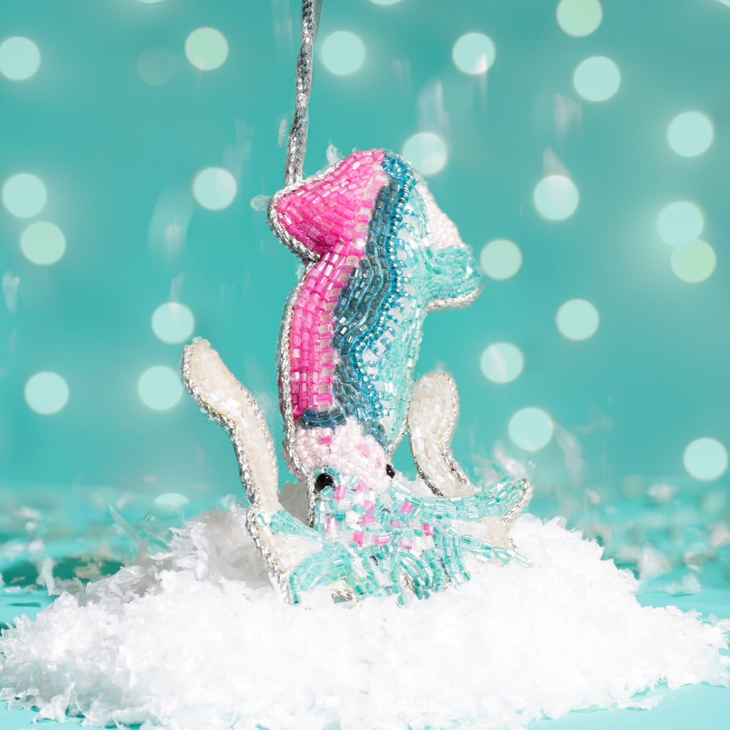 A colourful beaded squid decoration in pink, turquoise, and white tones, hanging above artificial snow with a teal background and soft bokeh lights.
