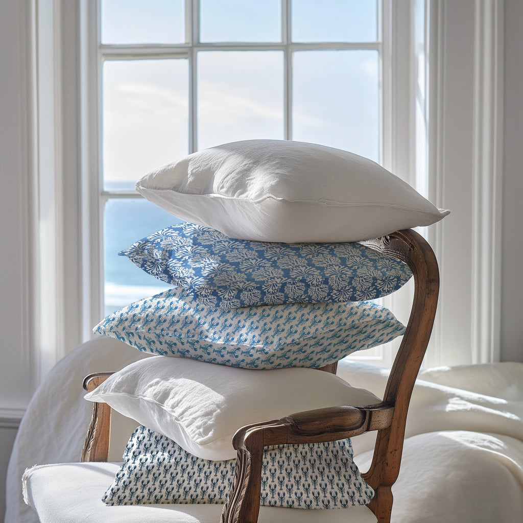 A stack of five cushions artfully balanced on a vintage wooden armchair in front of a large sunlit window with a view of the ocean. The cushions feature a mix of plain white and blue coastal-inspired patterns, including delicate coral and sea life motifs. Soft natural light fills the room, creating a serene and airy atmosphere.
