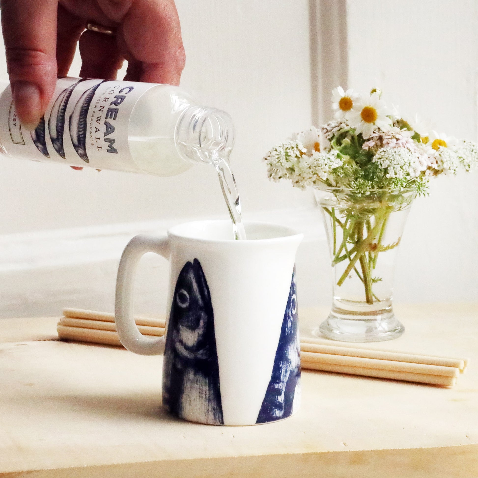 Mini Jug with mackerel heads design on with reeds and small posy of flowers behind with a bottle of Stargazey room diffuser oil being poured into the jug.