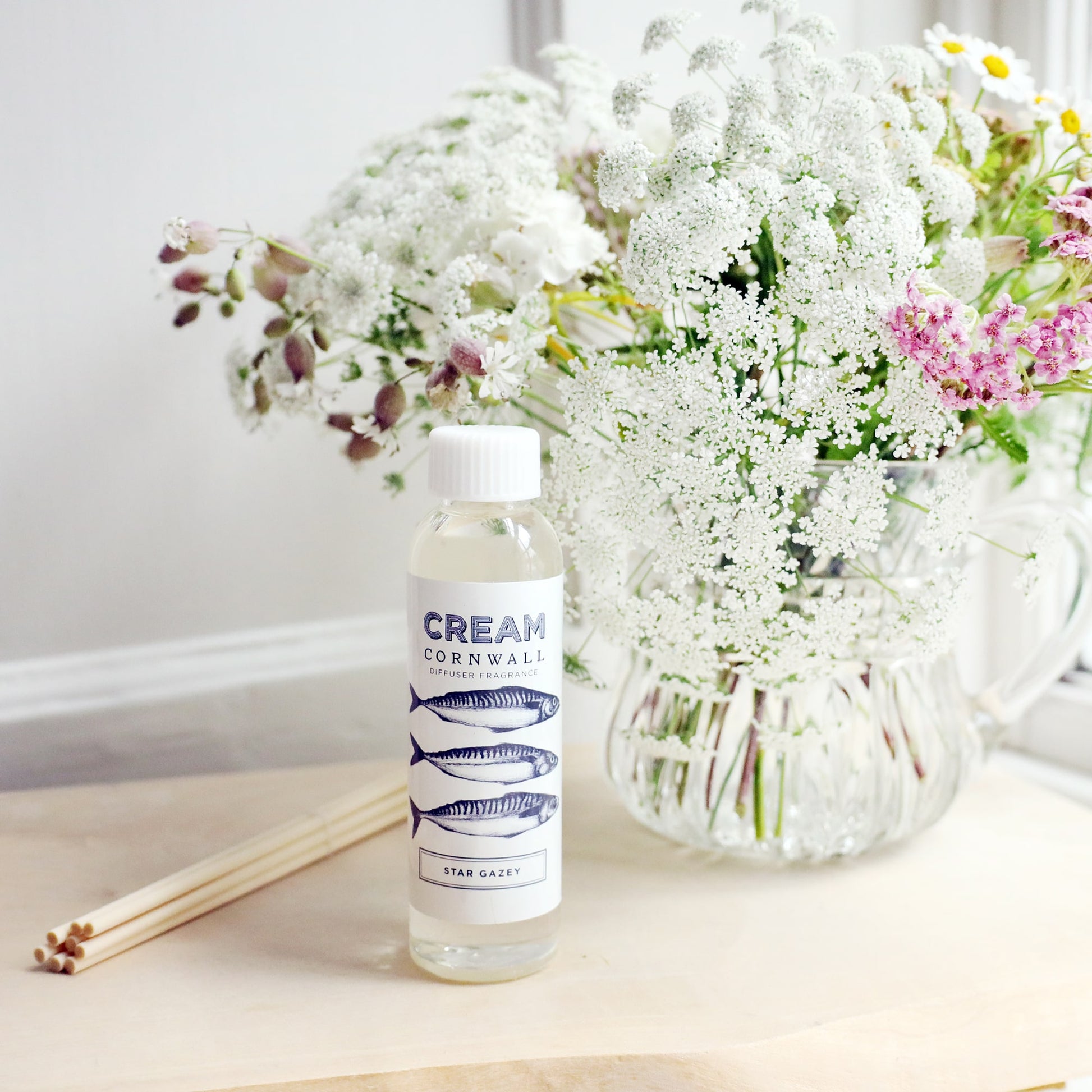 Bottle Stargazey of room diffuser oil with 3 mackerel design on the label, sitting on a wooden block with the diffuser reeds laying jut to the side, and a vase of soft coloured flowers sitting behind.