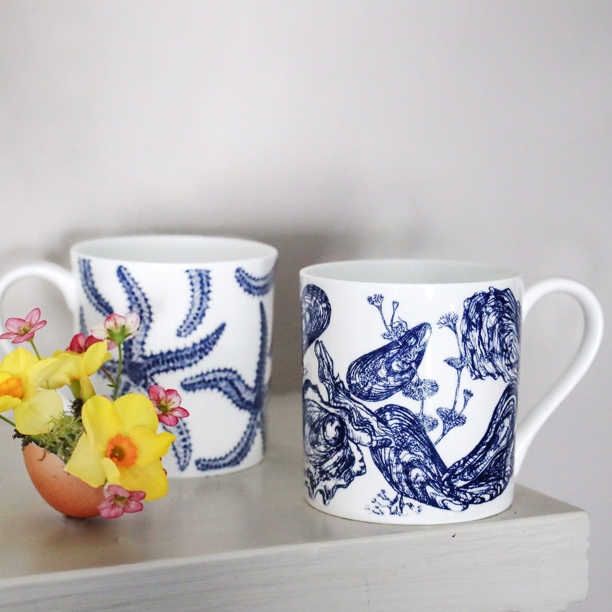 Two white mugs with blue starfish & mussel patterns on a shelf with flowers.