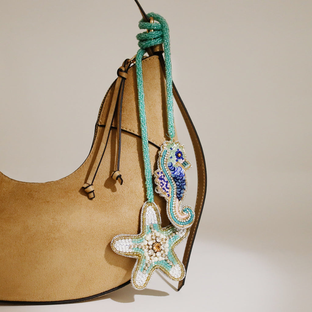 Beaded seahorse and starfish bag charms in turquoise, blue, and gold tones hanging from a tan suede handbag, with a knotted turquoise beaded cord.