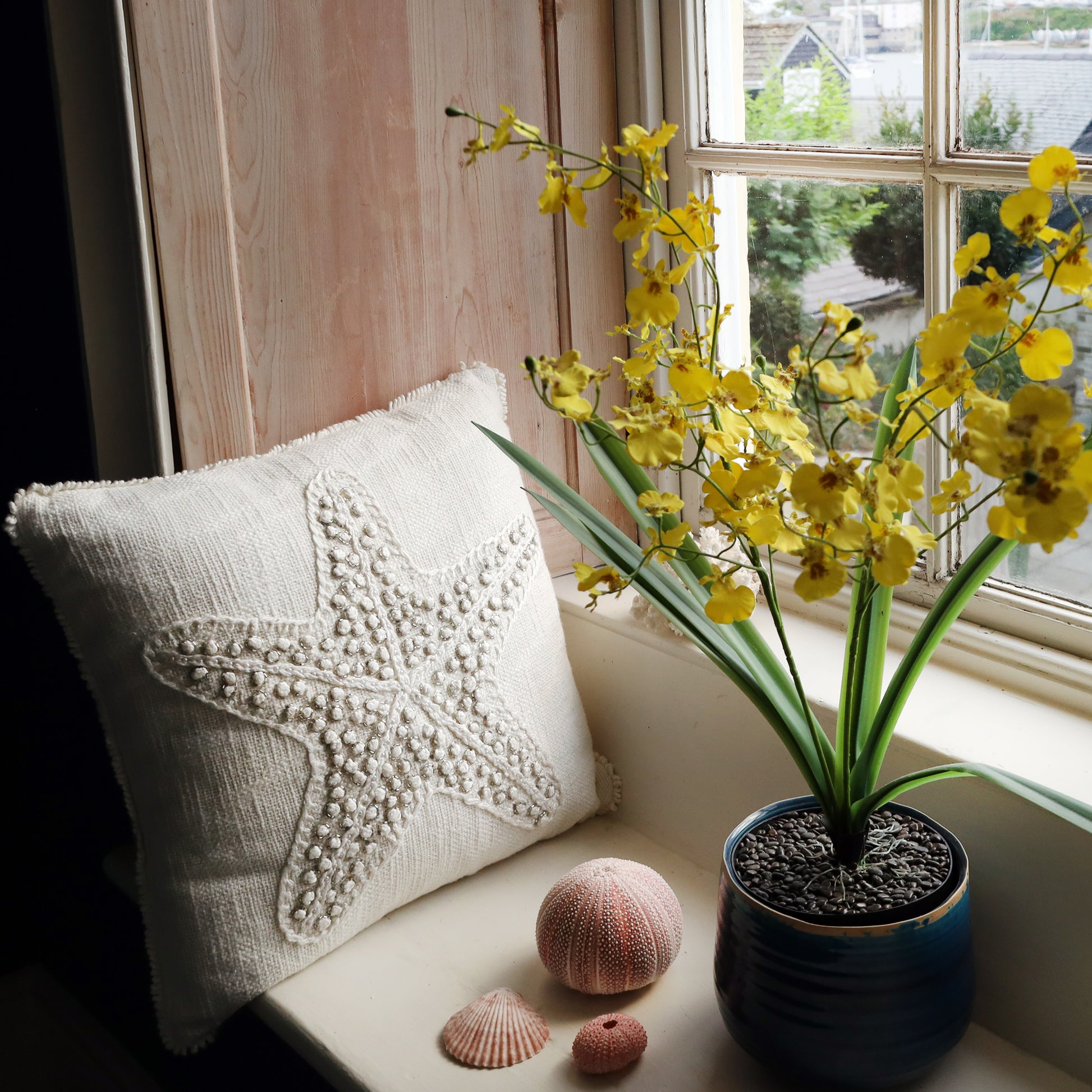 Decorative pillow with textured surface, potted yellow flowers, and seashells on a windowsill.