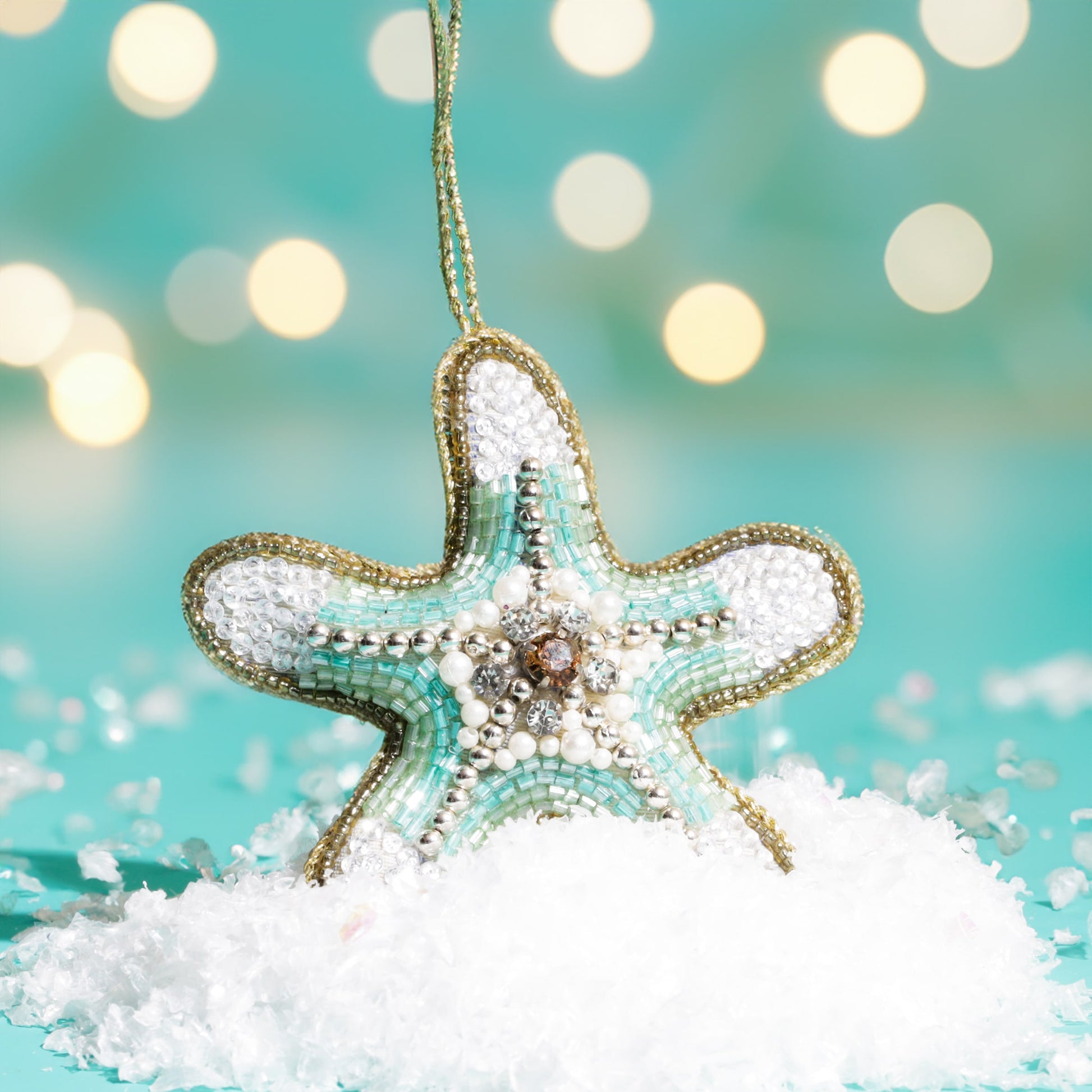 A turquoise and white beaded starfish decoration with pearl and crystal details, hanging above artificial snow with a teal background and softly glowing lights.