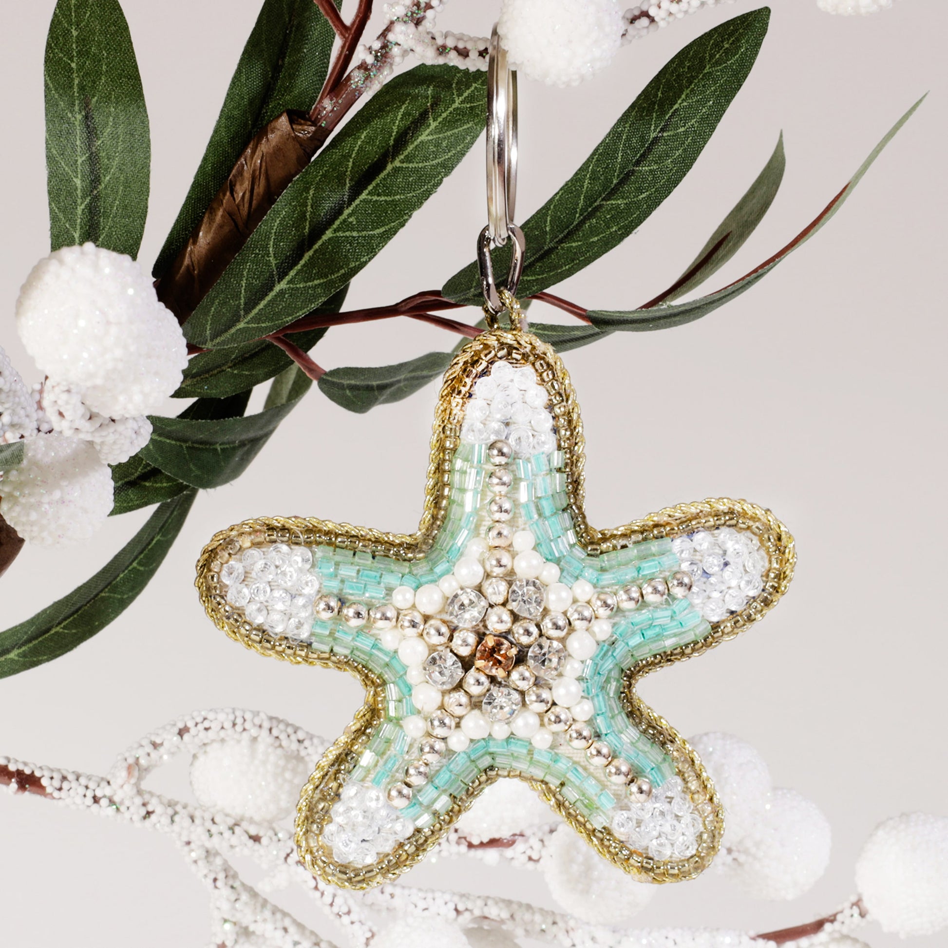 A beaded starfish keyring with turquoise, white, and gold beads hanging from a branch decorated with frosted white berries and green leaves.
