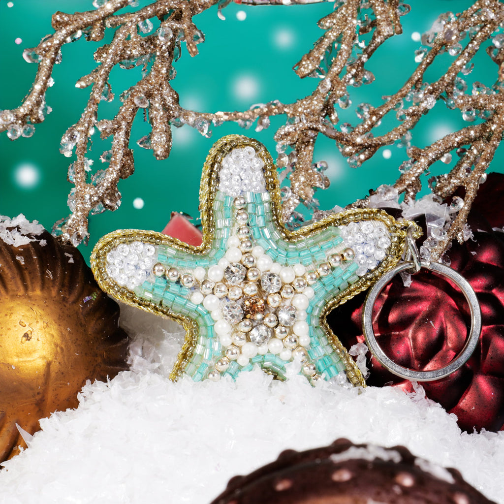 A beaded starfish keyring with turquoise, white, and gold detailing nestled in artificial snow among red and gold Christmas baubles and glittering gold branches against a teal background.