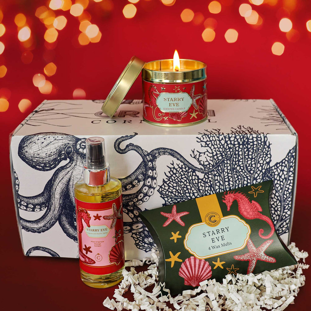 Starry Eve Christmas gift hamper featuring a lit scented candle, fragranced room spray and wax melts, displayed with a coastal-inspired gift box, white shredded paper and warm golden bokeh lights against a festive red background.