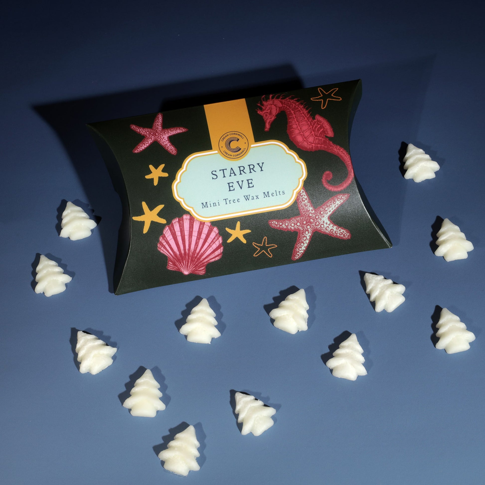 A flat lay image of a decorative package labeled "Starry Eve Mini Tree Wax Melts," featuring ocean-inspired illustrations of a pink seahorse, starfish, and seashells on a dark green background. Scattered around the package are small, white, tree-shaped wax melts, arranged neatly on a blue surface. The festive and coastal theme gives a whimsical, holiday feel to the scene.