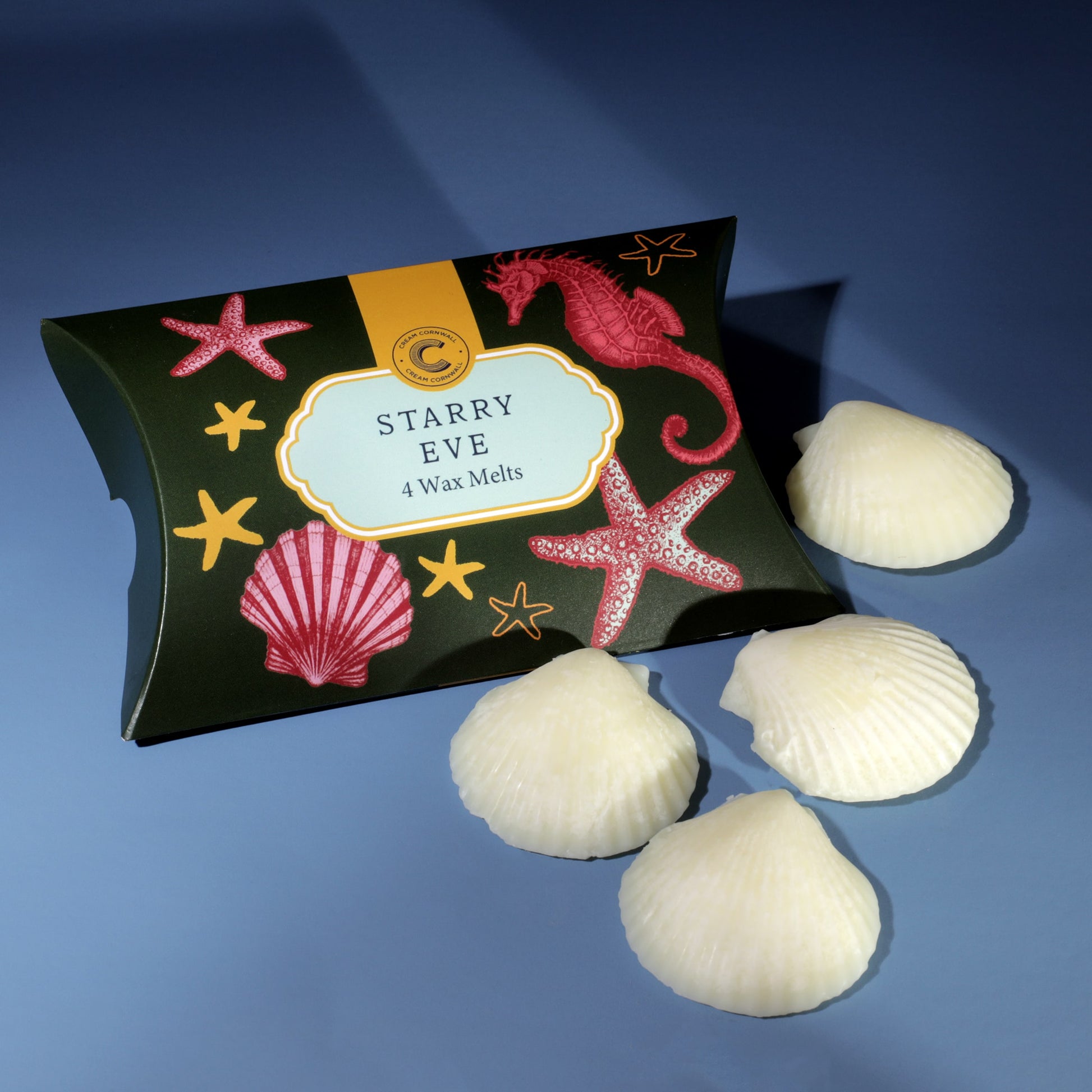 A box of Cream Cornwall’s Starry Eve wax melts, featuring a dark green box decorated with red seashells, starfish, and a seahorse design, displayed with four shell-shaped white wax melts on a blue surface.