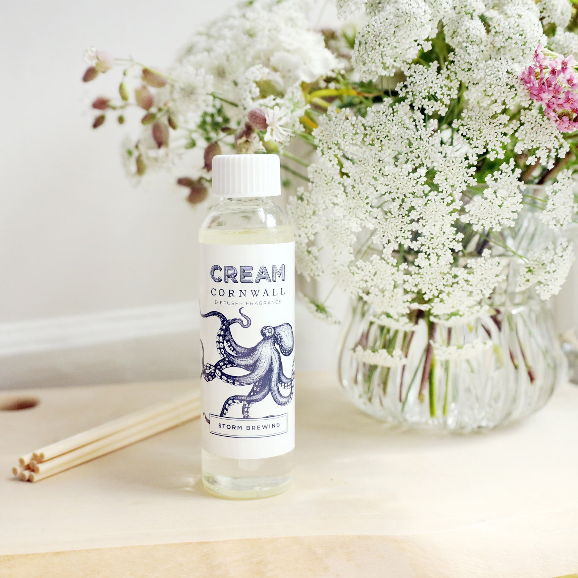 Bottle of Storm Brewing room diffuser oil with octopus design on the label, sitting on a wooden block with the diffuser reeds laying jut to the side, and a vase of soft coloured flowers sitting behind.