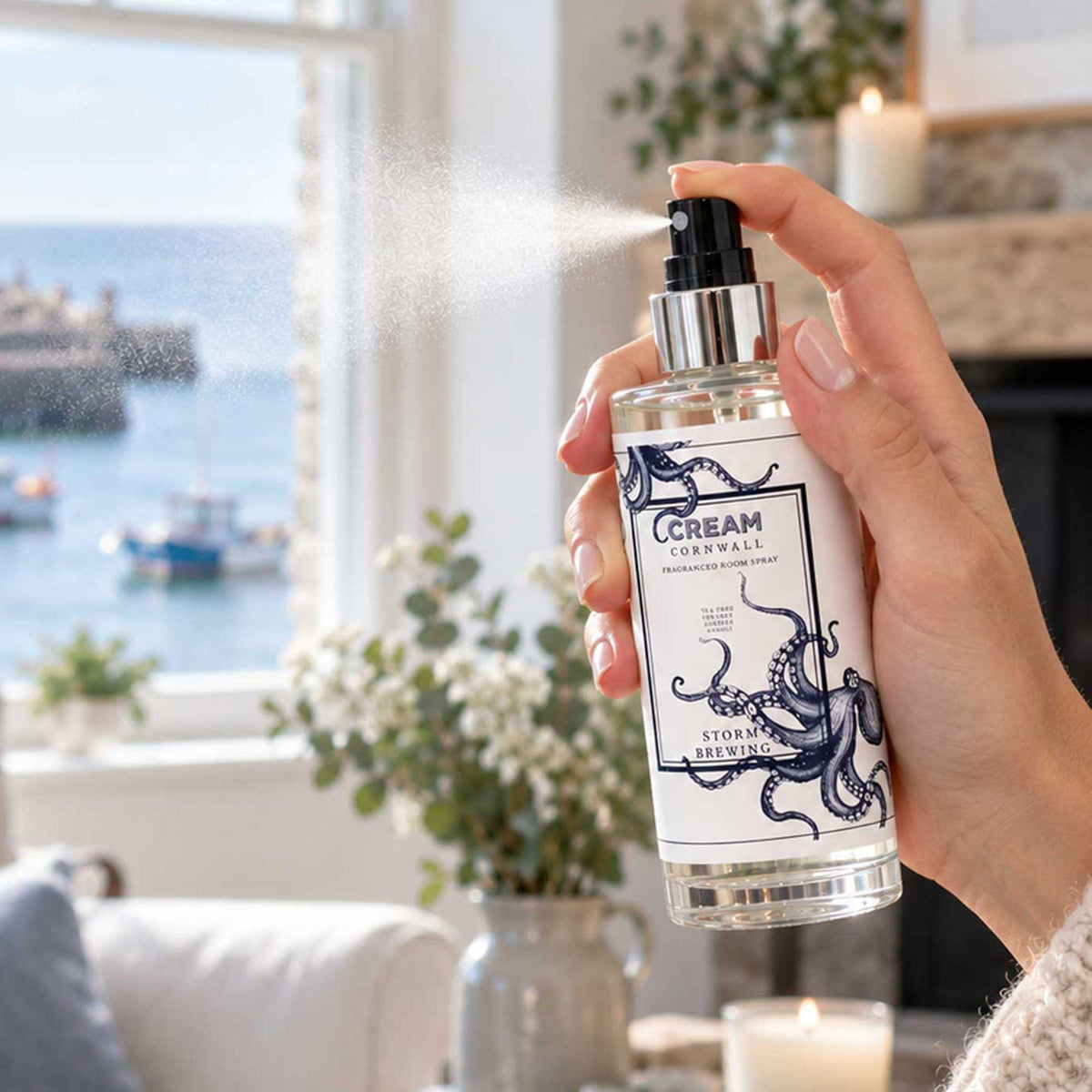 Hand spraying Cream Cornwall Storm Brewing fragranced room spray, featuring a navy octopus design, in a cosy coastal living room with fresh flowers, lit candles and a harbour view through the window.