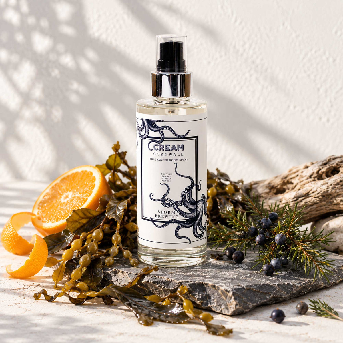 Cream Cornwall Storm Brewing fragranced room spray with navy octopus design, displayed on natural stone alongside fresh orange, juniper berries, seaweed and driftwood against a softly sunlit neutral background.