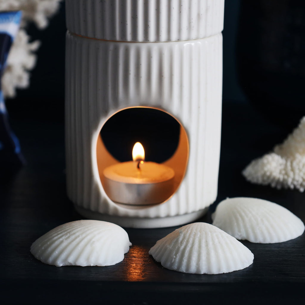 White ceramic lantern with a lit candle inside, surrounded by seashells on a dark surface.