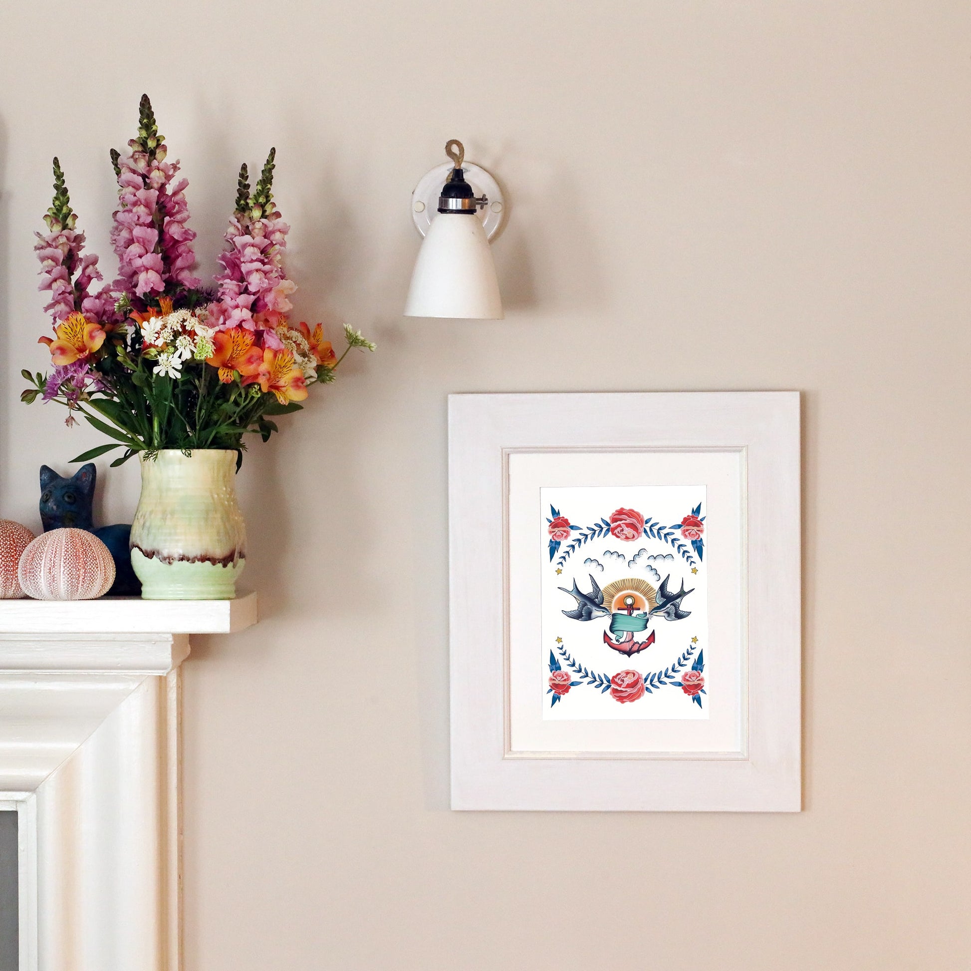 A framed nautical-style print featuring two swallows, an anchor, and roses hangs on a pale wall beneath a vintage-style wall lamp. A mantel nearby holds a vase of colorful flowers, decorative seashells, and a ceramic cat figurine.