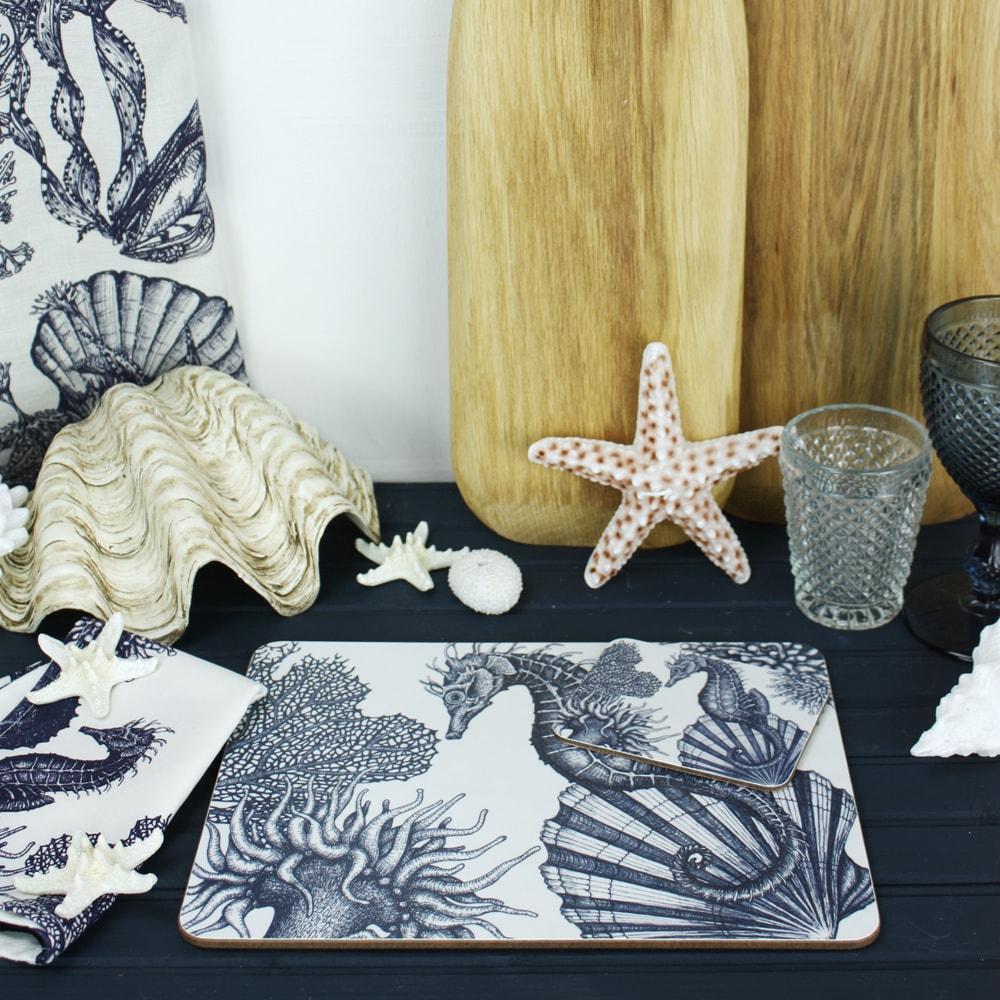 Seahorse Design in Navy on a white Coaster with a matching Placemat.On the table are decorative shells and a matching napkin