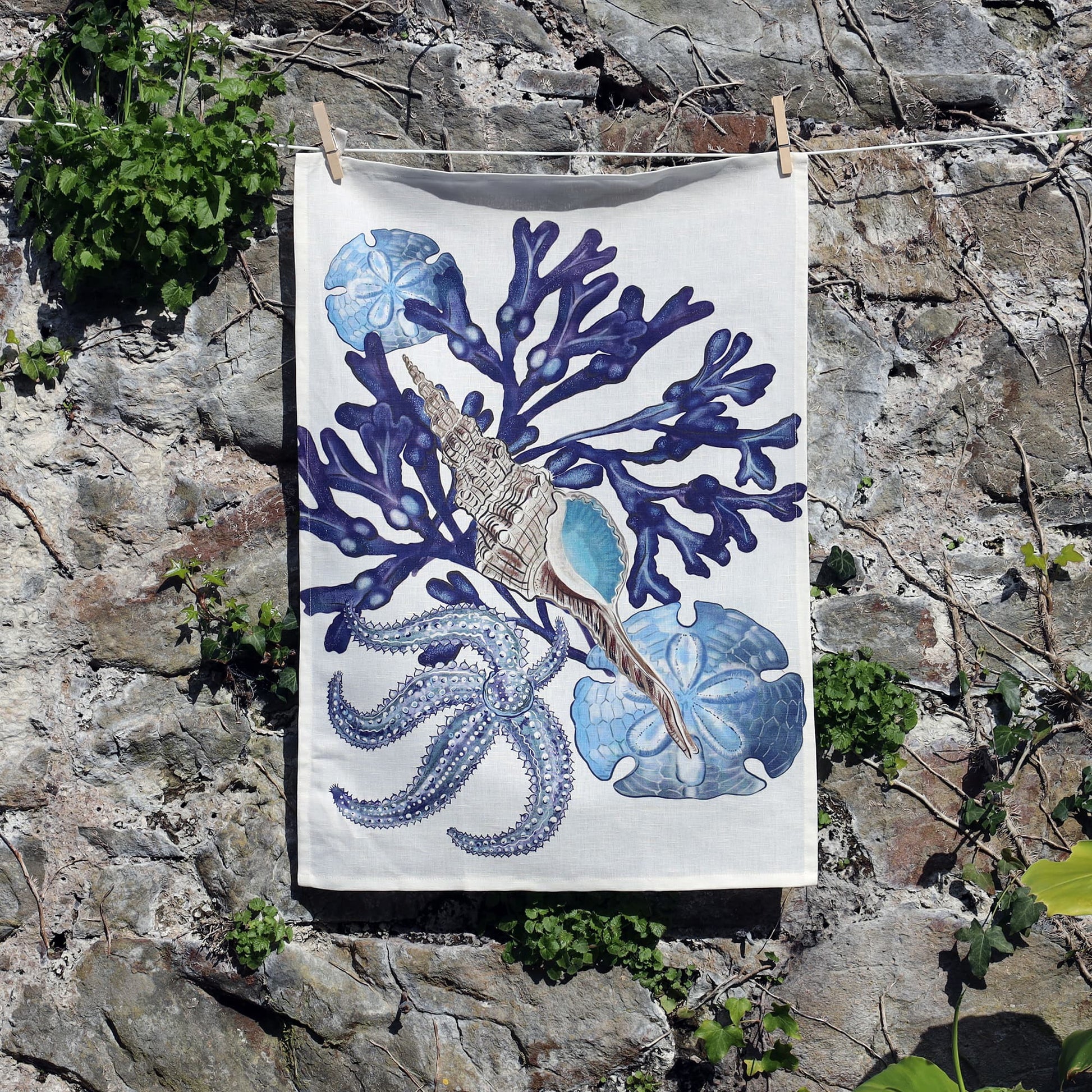 A coastal-themed tea towel hangs on a clothesline against a weathered stone wall. The design features rich blue coral branches, a detailed spiral seashell with turquoise interior, an elegant octopus with textured tentacles, and blue sand dollar motifs. Small patches of green ivy grow along the wall, adding a natural contrast to the intricate marine illustration on the tea towel.