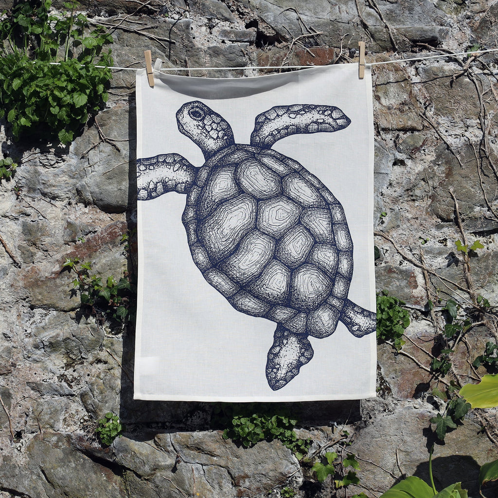 A tea towel with an intricate navy blue line drawing of a sea turtle, pegged to a clothesline and hanging against a stone wall with green foliage growing around it. The towel features detailed textures on the turtle's shell and flippers.