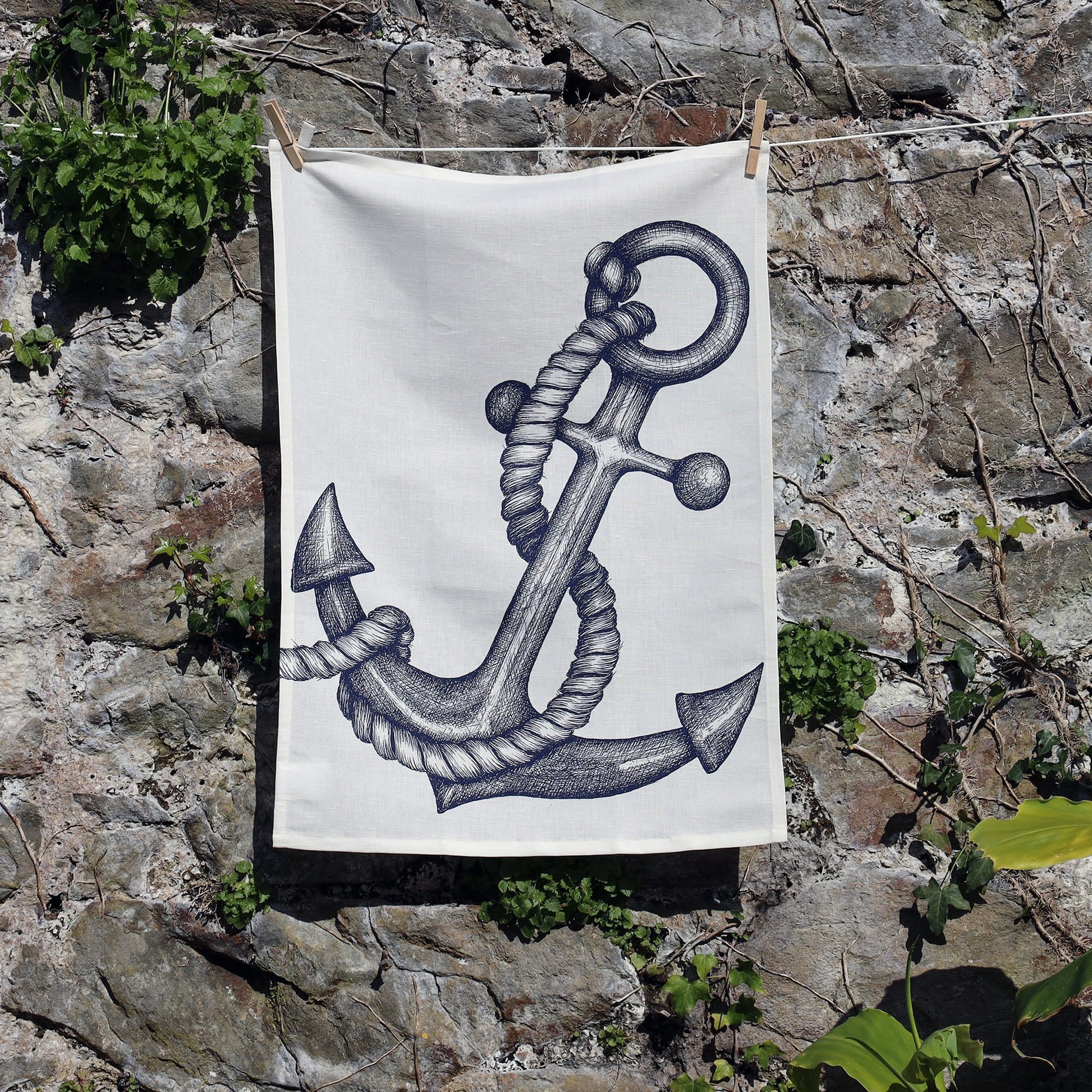 A tea towel featuring a detailed black ink illustration of an anchor wrapped in thick rope hangs on a clothesline against a rugged stone wall. Green vines and foliage grow around the rocks, adding a natural contrast to the nautical-themed design.