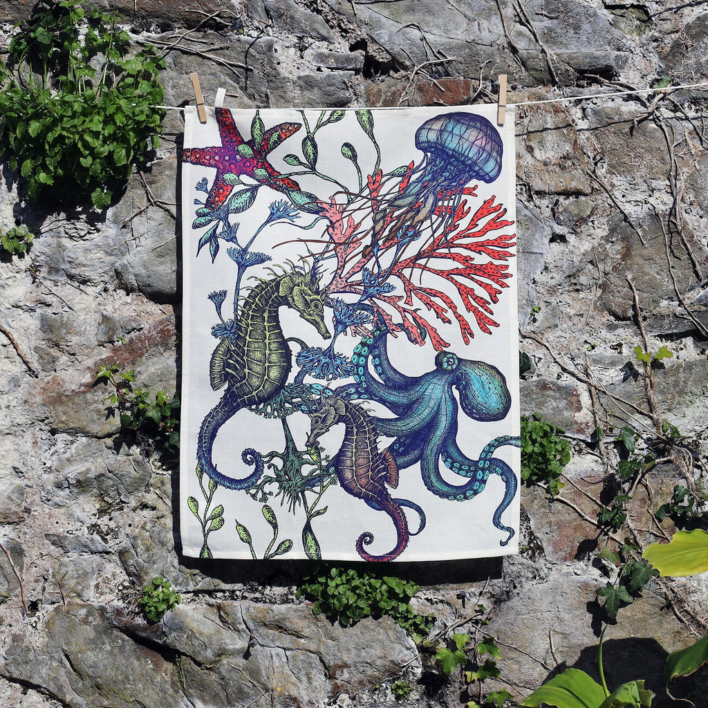 A tea towel hangs against a stone wall, featuring a vibrant underwater illustration with seahorses, an octopus, jellyfish, starfish, and colorful coral and seaweed in bold shades of blue, green, red, and purple. The lively design contrasts with the rustic natural background, highlighting the marine-themed artwork.