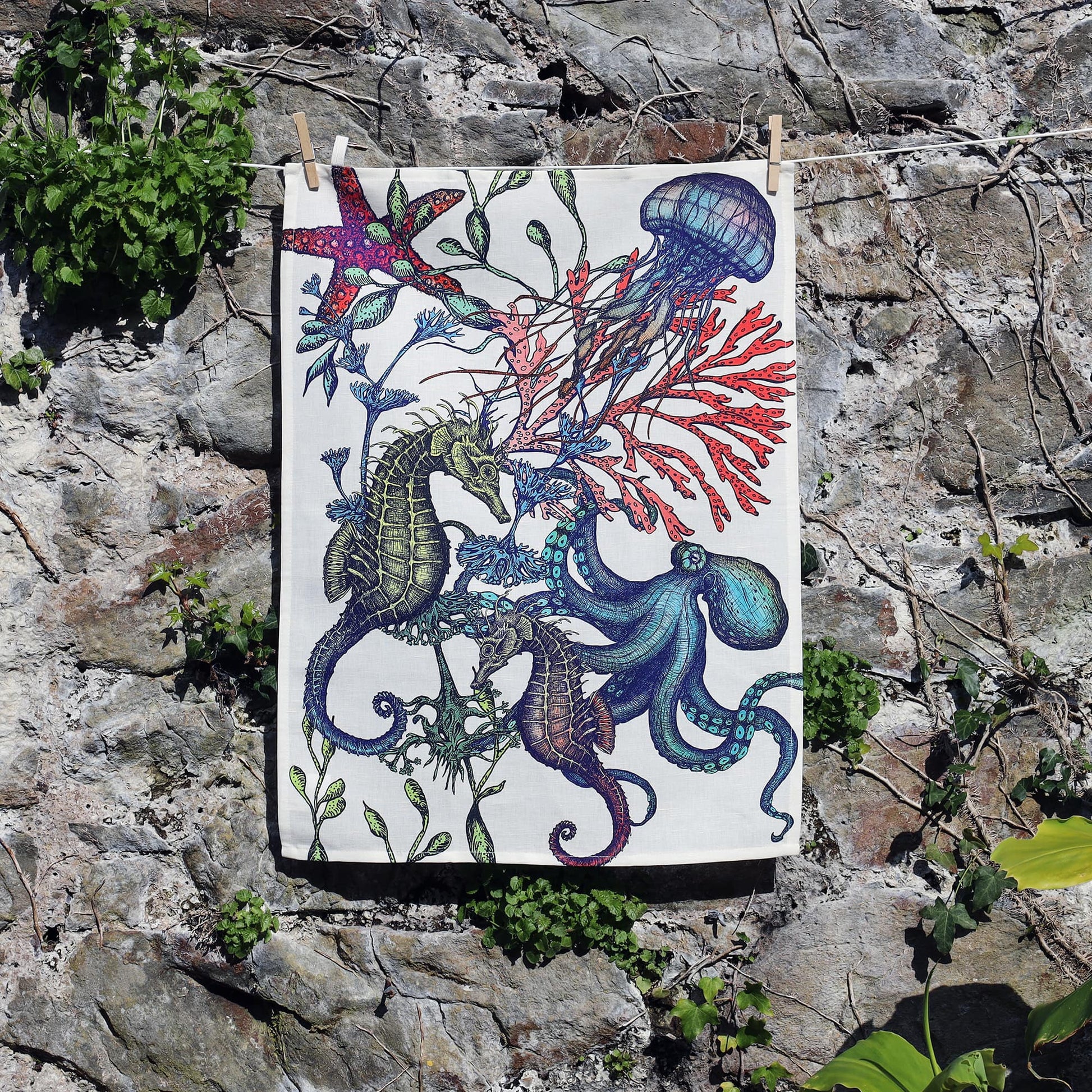 A tea towel hangs against a stone wall, featuring a vibrant underwater illustration with seahorses, an octopus, jellyfish, starfish, and colorful coral and seaweed in bold shades of blue, green, red, and purple. The lively design contrasts with the rustic natural background, highlighting the marine-themed artwork.