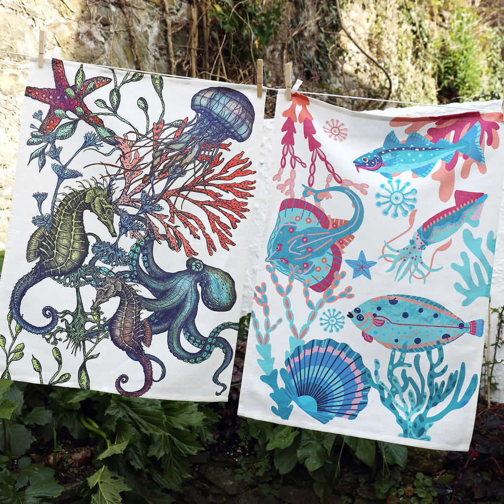 Two vibrant tea towels hang side by side on a washing line against a garden stone wall. The left towel features a detailed illustration of seahorses, an octopus, jellyfish, and red coral in deep, earthy tones. The right towel displays a playful, colorful underwater scene with a squid, ray, fish, and sea plants in bright turquoise, pink, and coral hues.