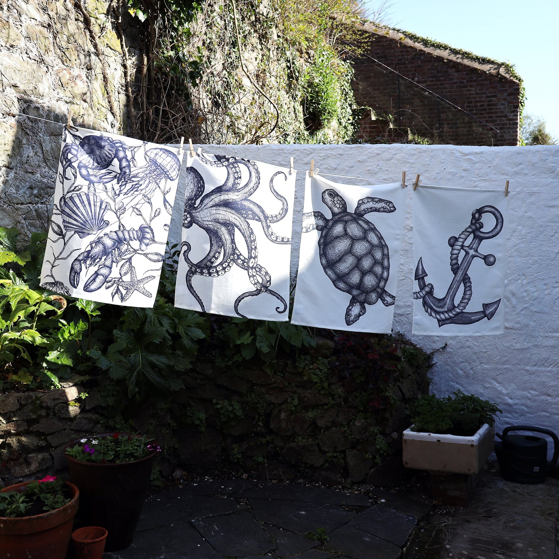 Four monochrome tea towels hang side by side on a washing line in a sunlit garden courtyard. From left to right, the designs feature: a lobster, jellyfish, and seashells with seaweed; a large octopus with swirling tentacles; a detailed sea turtle; and a nautical anchor wrapped in rope. The scene is set against a stone wall and whitewashed building, with greenery and flower pots below.