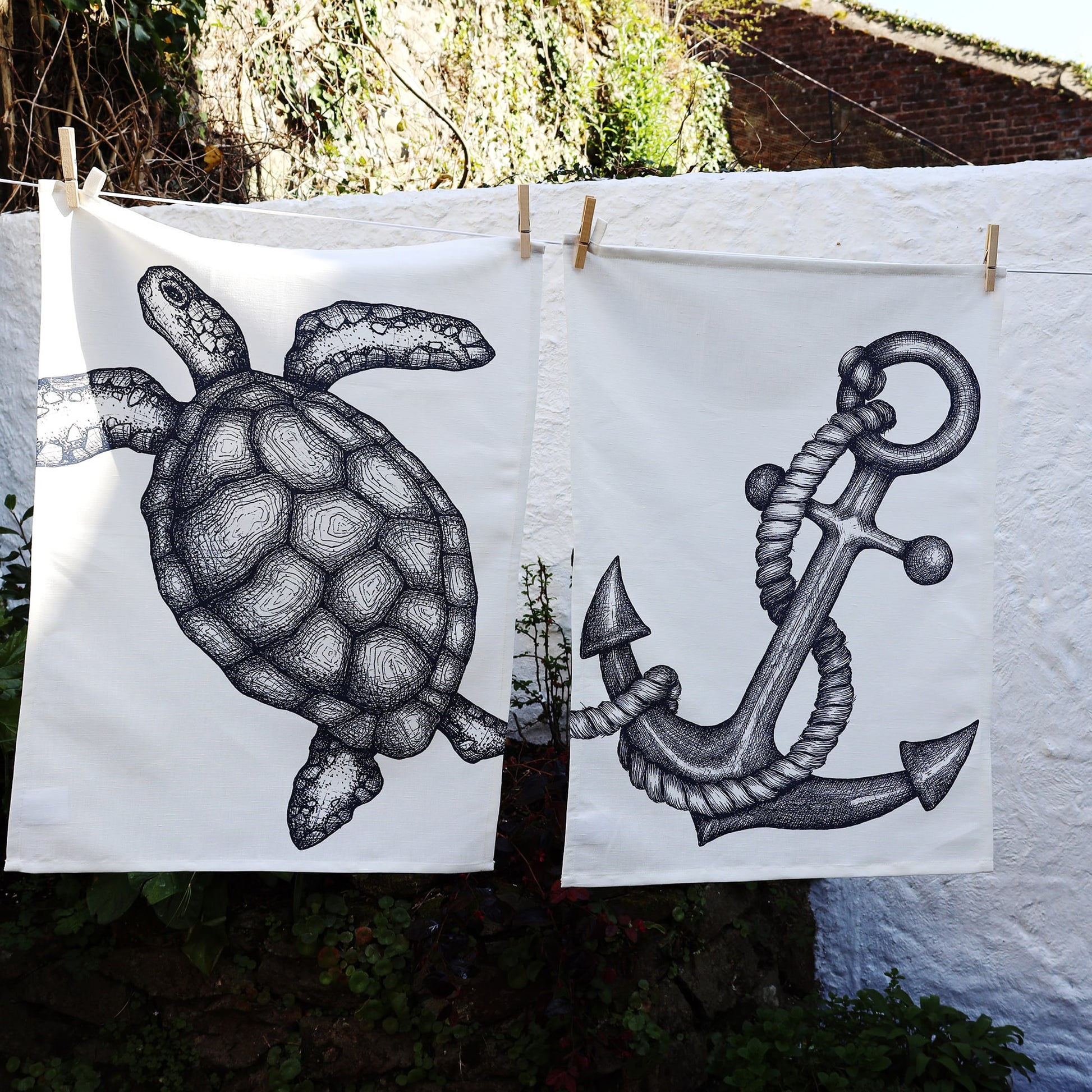 Two nautical-themed tea towels hang side by side on a washing line against a white stucco wall. The towel on the left features a finely detailed illustration of a sea turtle, while the towel on the right displays a bold anchor wrapped in thick rope. Sunlight casts shadows on the wall, adding depth to the serene coastal garden setting.
