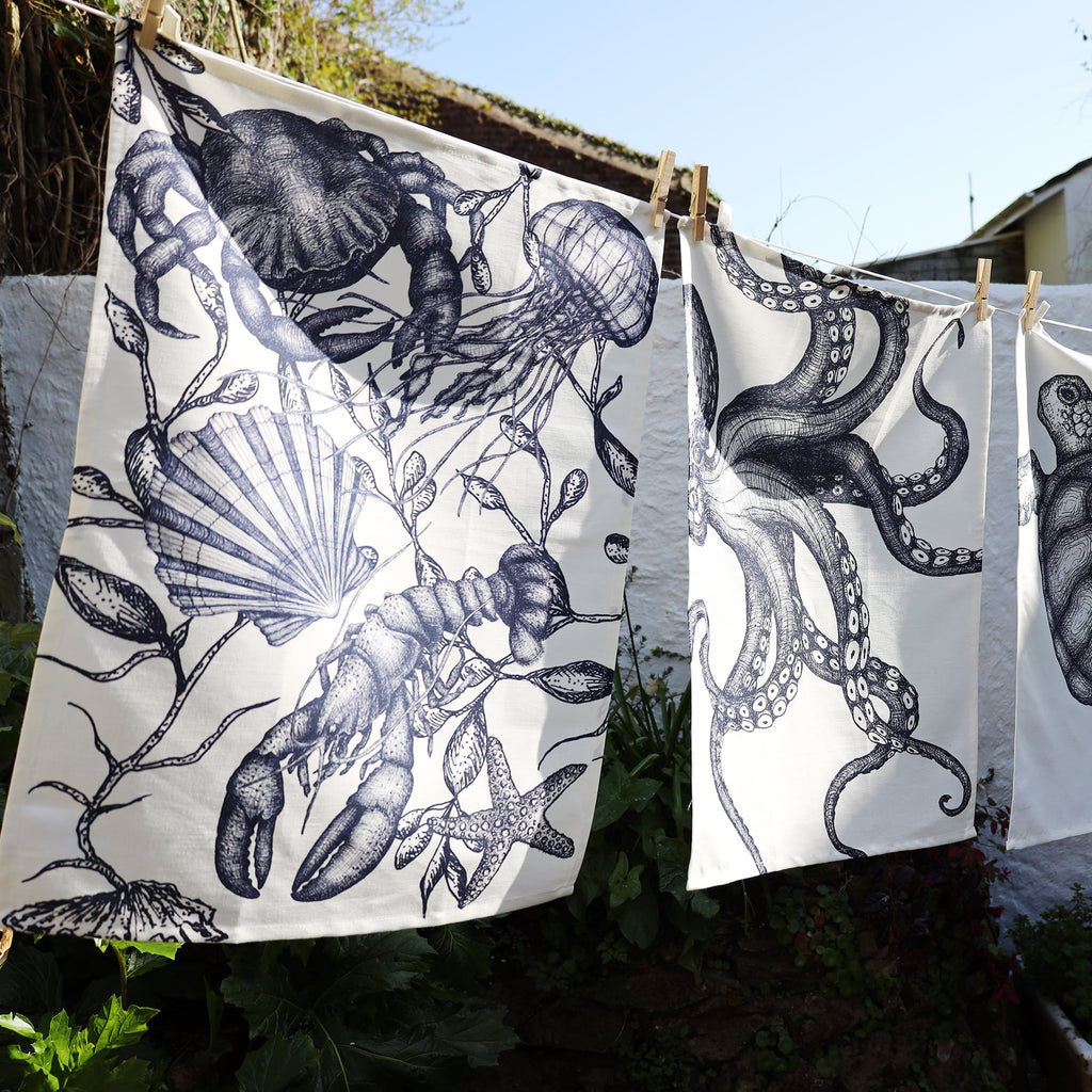 Three nautical-themed tea towels hang on a washing line in the sunlight, secured with wooden pegs. The left towel features detailed monochrome illustrations of a lobster, jellyfish, scallop shell, crab, seaweed, and starfish. The middle towel showcases a close-up of an intricately drawn octopus with swirling tentacles, while the right towel partially reveals a continuation of the oceanic theme. The background includes a white wall, a garden with lush greenery, and a clear blue sky.