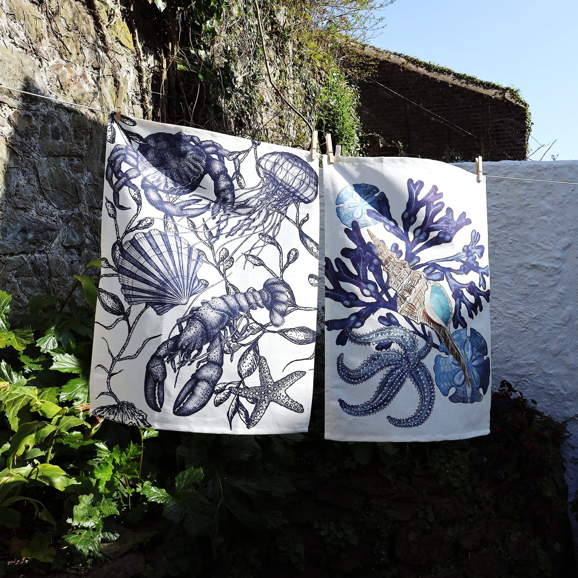 Two coastal-themed tea towels hang on a clothesline outdoors against a backdrop of a stone wall and greenery. The towel on the left features intricate navy blue illustrations of a lobster, jellyfish, seashells, and starfish on a white background. The towel on the right displays a stylized underwater scene with deep blue coral, a detailed seashell, and a swirling octopus. The towels are bathed in natural sunlight, highlighting their bold marine-inspired designs.