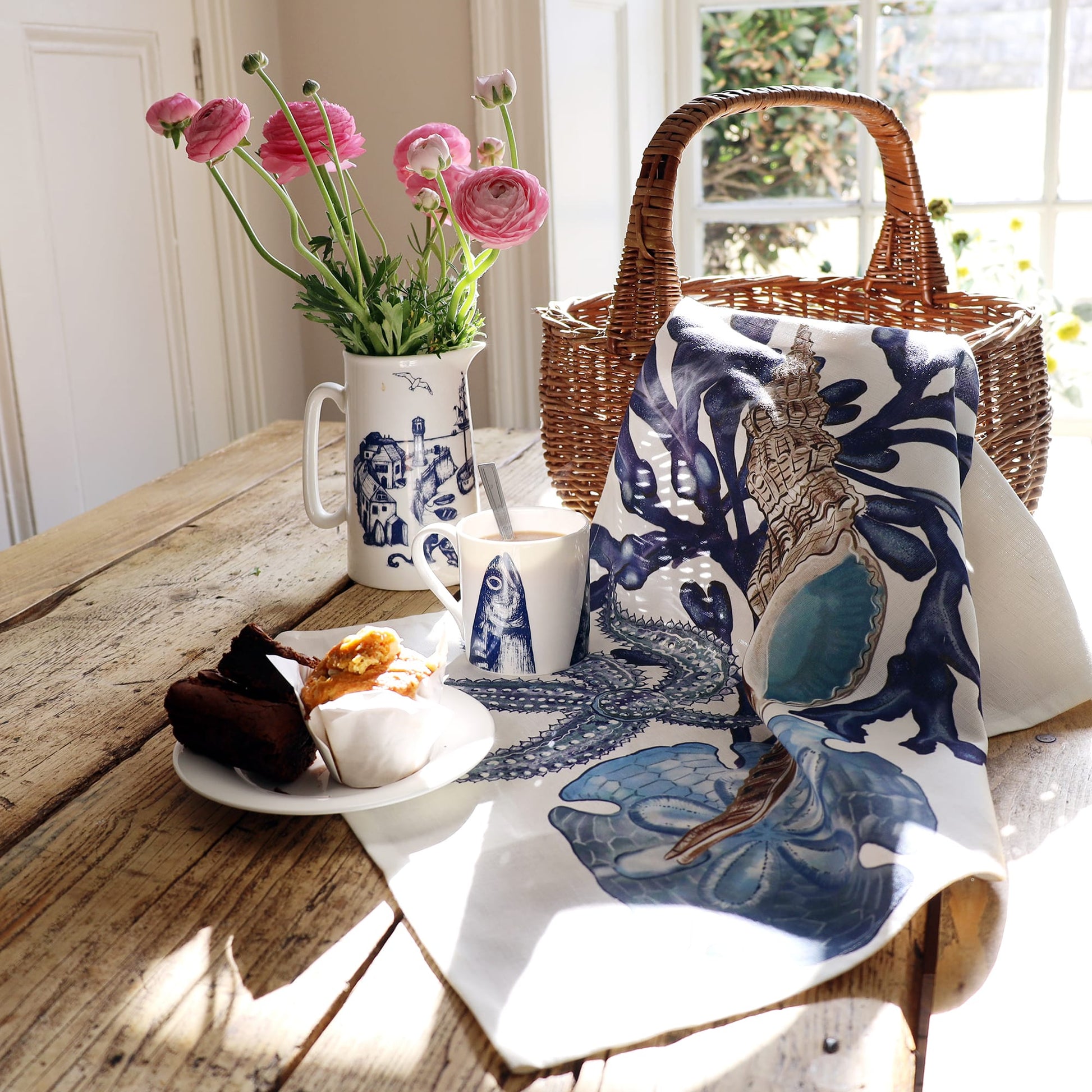 A rustic wooden table set by a sunny window displays a coastal-themed scene featuring a wicker basket, a white tea towel with a bold blue and brown seashell and coral illustration, and a matching ceramic jug and mug adorned with nautical drawings. A plate with pastries and brownies, a spoon in the mug, and a vase with pink ranunculus flowers complete the cozy and seaside-inspired setting.