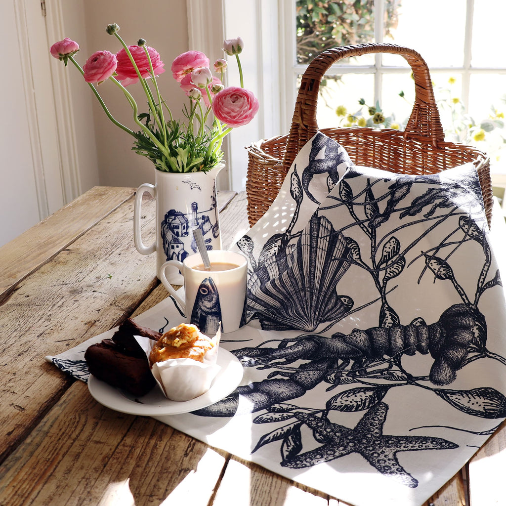 A coastal-themed table setting featuring a tea towel with intricate navy blue illustrations of a lobster, starfish, seaweed, and shell draped over a rustic wooden table. The setup includes a wicker basket, a white mug and jug with nautical blue artwork, a plate with a muffin and brownies, and a vase of fresh pink ranunculus flowers. Sunlight pours in through a nearby window, casting soft shadows and enhancing the seaside-inspired atmosphere.