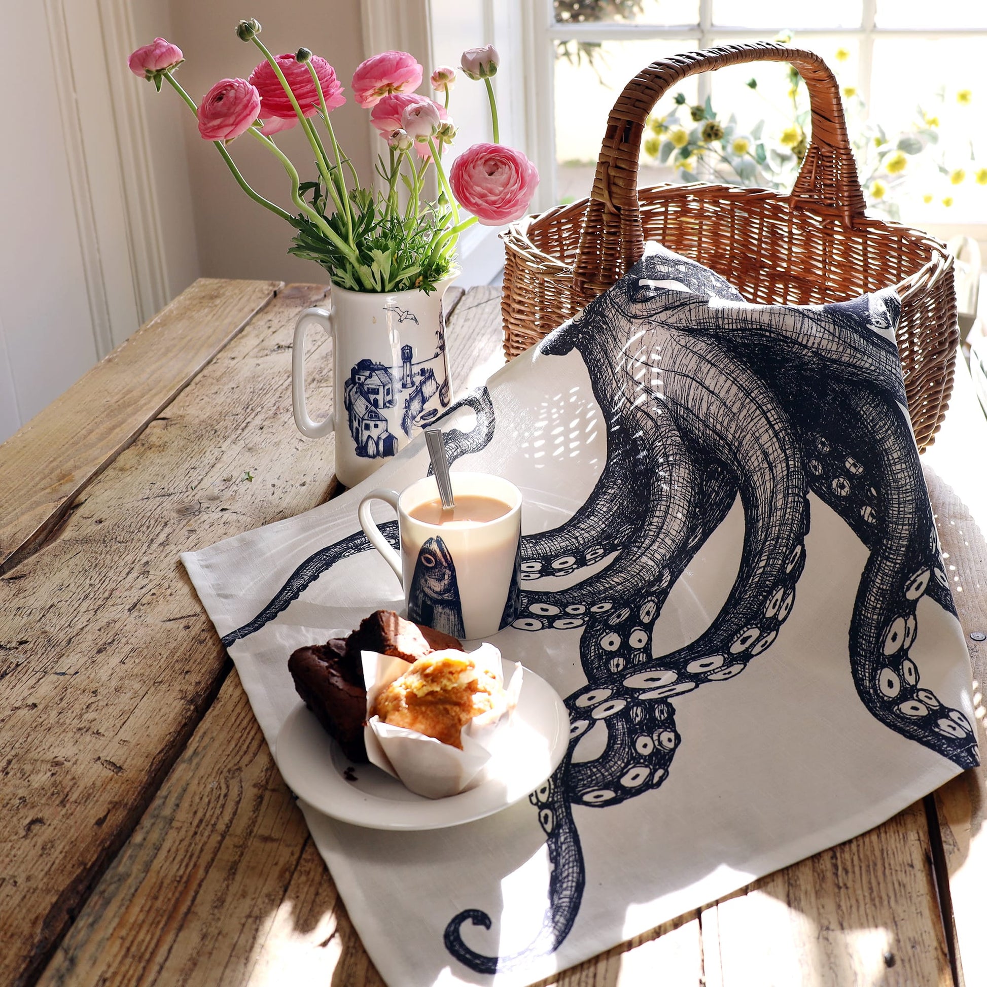 A rustic wooden table is set with a nautical-themed tea towel featuring a detailed navy octopus illustration. On top of the towel sits a plate with a muffin and chocolate cake slices, a white mug with a fish design filled with tea, and a spoon. A wicker basket and a jug of pink ranunculus flowers sit in the background near a sunlit window, creating a cozy, seaside-inspired breakfast scene.