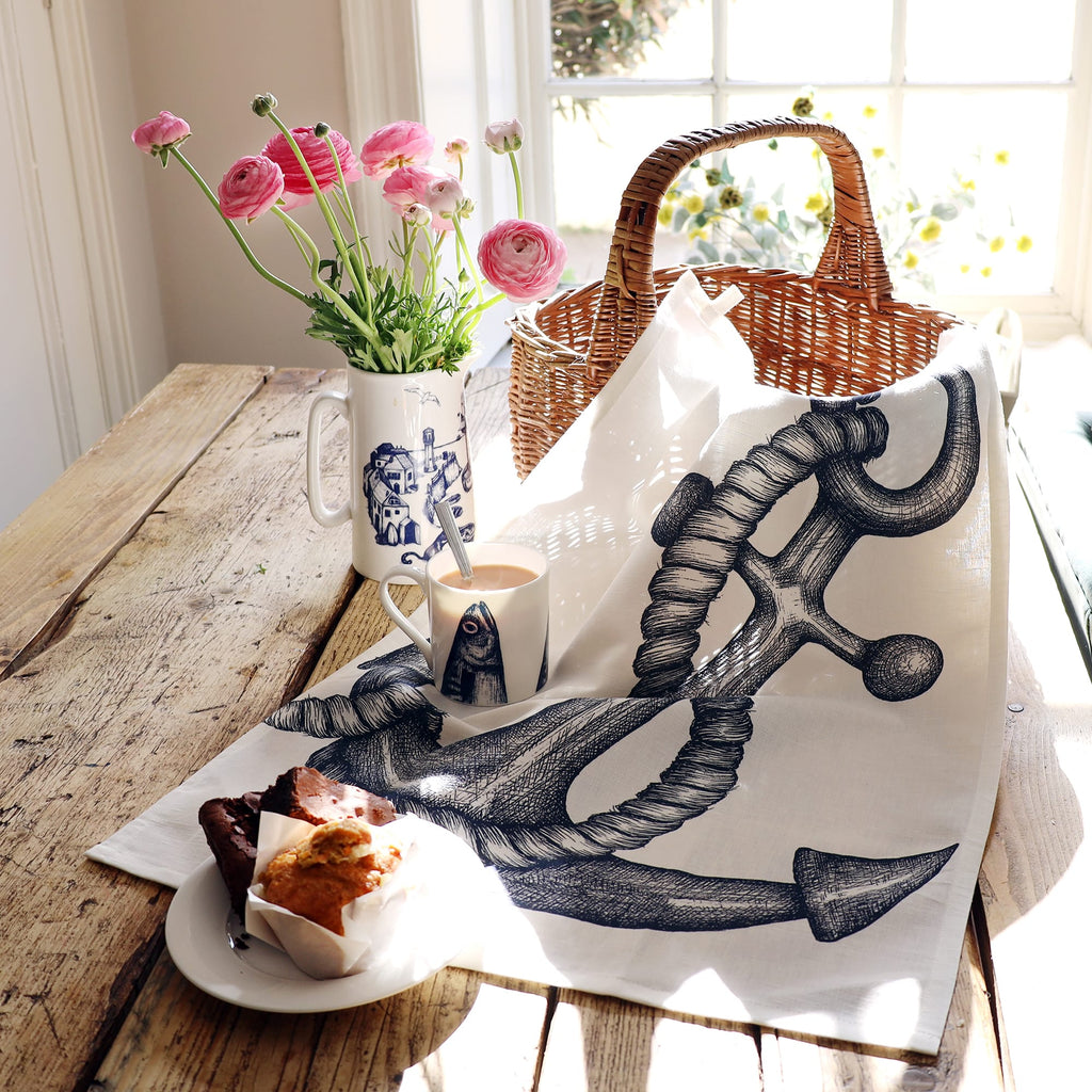 A rustic wooden table set by a sunny window features a nautical-themed tea towel with a large black ink illustration of an anchor wrapped in rope. A wicker picnic basket, a plate of brownies and a muffin, a fish-illustrated mug with tea, and a tall jug holding fresh pink ranunculus flowers complete the cozy seaside-inspired scene.