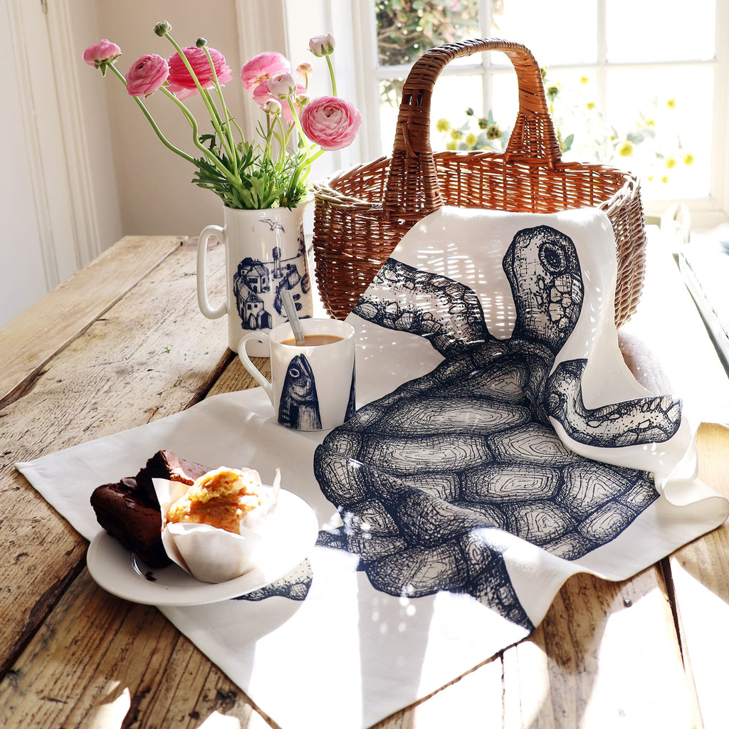 A tea towel with a detailed navy blue illustration of a sea turtle draped over a rustic wooden table beside a wicker basket. The scene includes a white mug with a fish design, a matching jug holding pink ranunculus flowers, and a plate with a chocolate brownie and a muffin, all bathed in warm sunlight streaming through a nearby window.