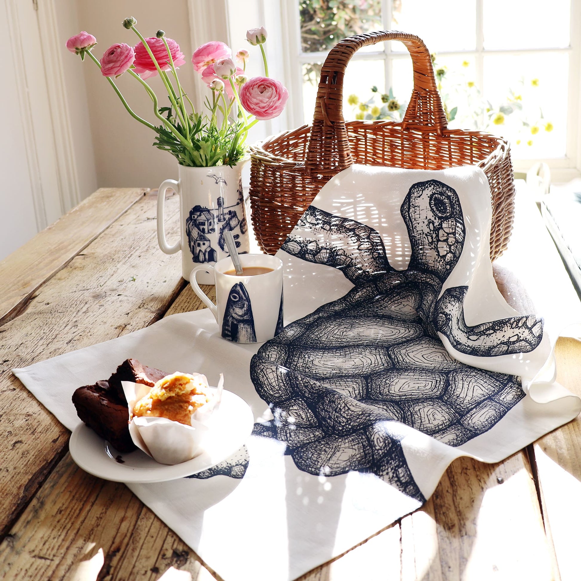 A tea towel with a detailed navy blue illustration of a sea turtle draped over a rustic wooden table beside a wicker basket. The scene includes a white mug with a fish design, a matching jug holding pink ranunculus flowers, and a plate with a chocolate brownie and a muffin, all bathed in warm sunlight streaming through a nearby window.
