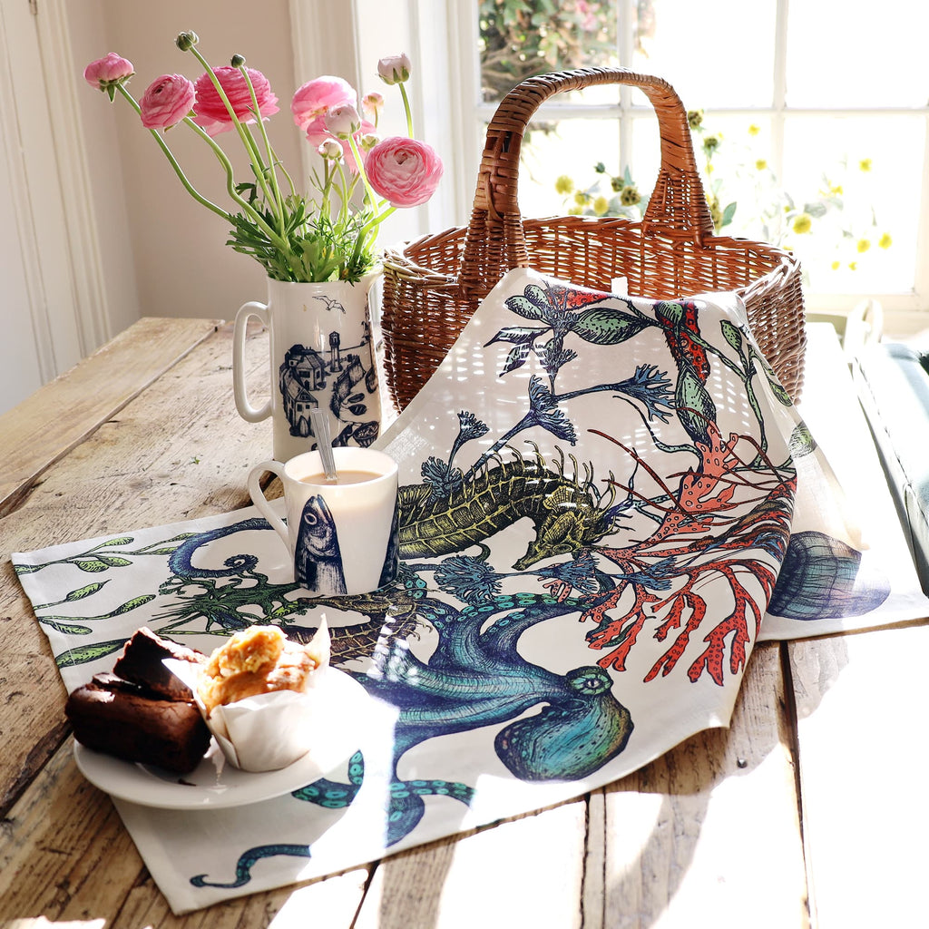 A tea towel with a vibrant underwater design featuring seahorses, an octopus, seaweed, and coral lies draped across a rustic wooden table. The table is set near a sunlit window with a wicker basket, a plate of baked goods, a mug of tea, and a tall jug holding pink ranunculus flowers, creating a warm and inviting seaside-themed breakfast scene.