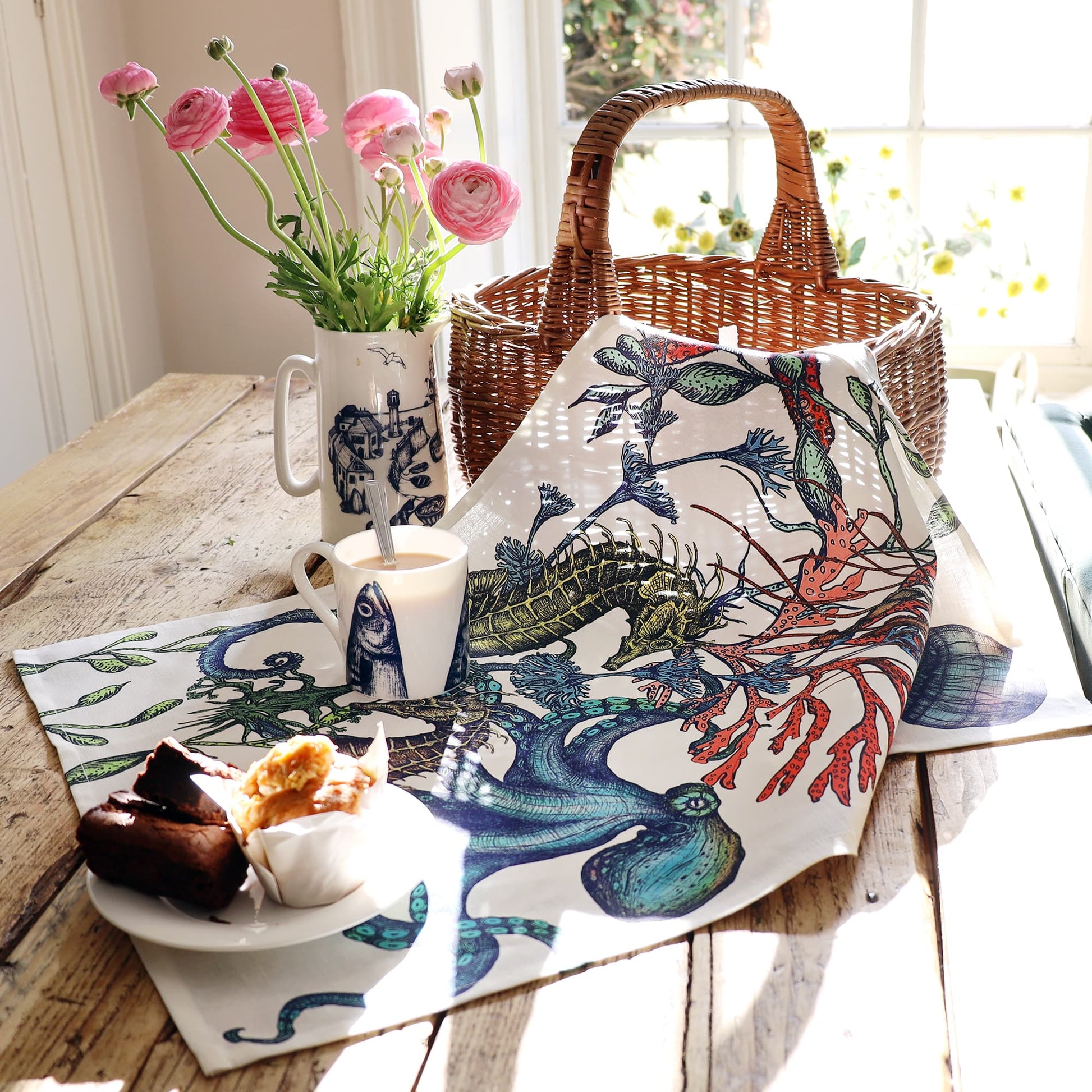 A tea towel with a vibrant underwater design featuring seahorses, an octopus, seaweed, and coral lies draped across a rustic wooden table. The table is set near a sunlit window with a wicker basket, a plate of baked goods, a mug of tea, and a tall jug holding pink ranunculus flowers, creating a warm and inviting seaside-themed breakfast scene.