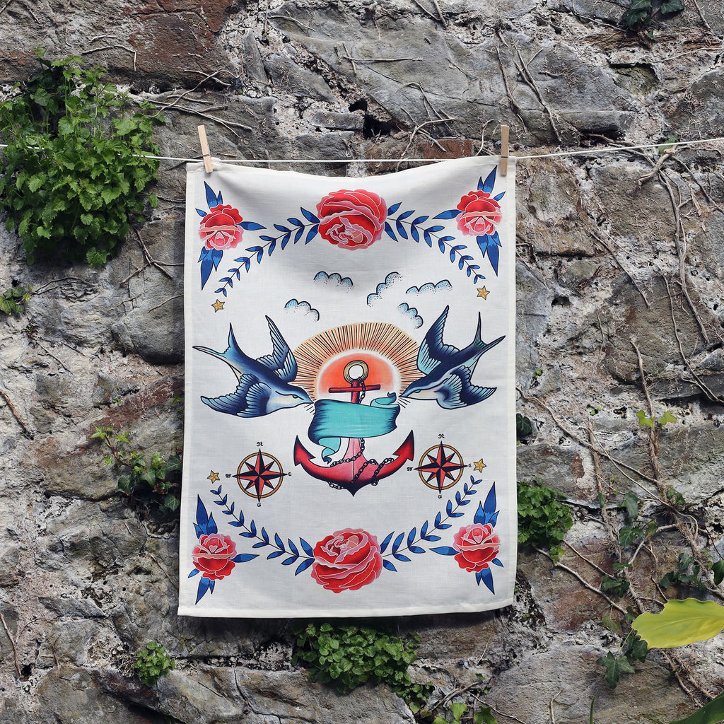 A tea towel hangs against a rustic stone wall, featuring a bold, vintage tattoo-inspired nautical design. At the center is a pink and red anchor with a blue banner, flanked by two flying blue swallows and surrounded by red roses, compass stars, and laurel-like leaf motifs. A stylized sun with rays and small clouds completes the symmetrical and colorful composition.