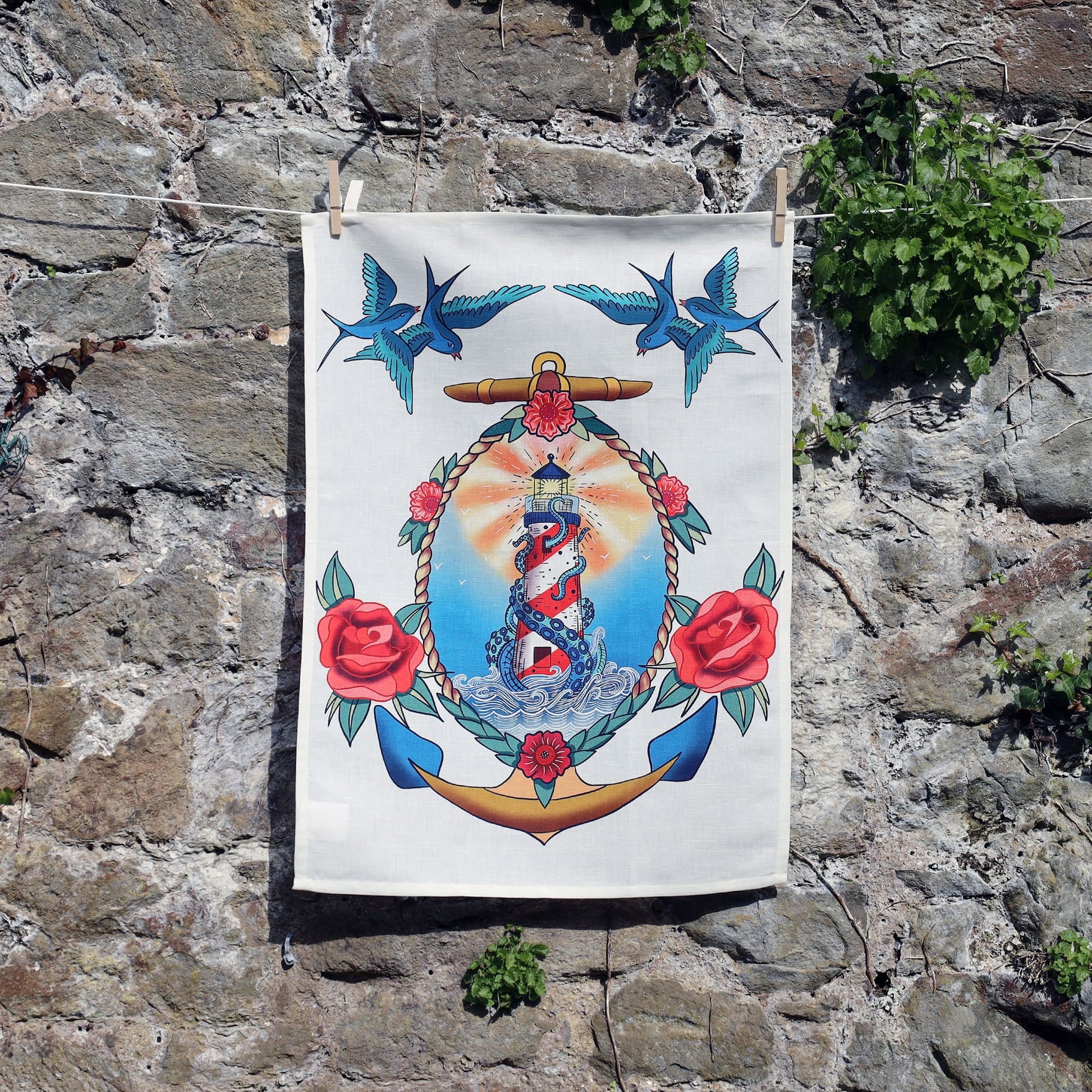 A tea towel hangs against a stone wall, featuring a vibrant nautical tattoo-inspired design. At the center is a red and white lighthouse surrounded by waves and a coiled blue sea serpent. The lighthouse is framed by an anchor, rope, and red roses, with three blue swallows flying overhead. The design is colorful and symmetrical, with a classic vintage tattoo style.